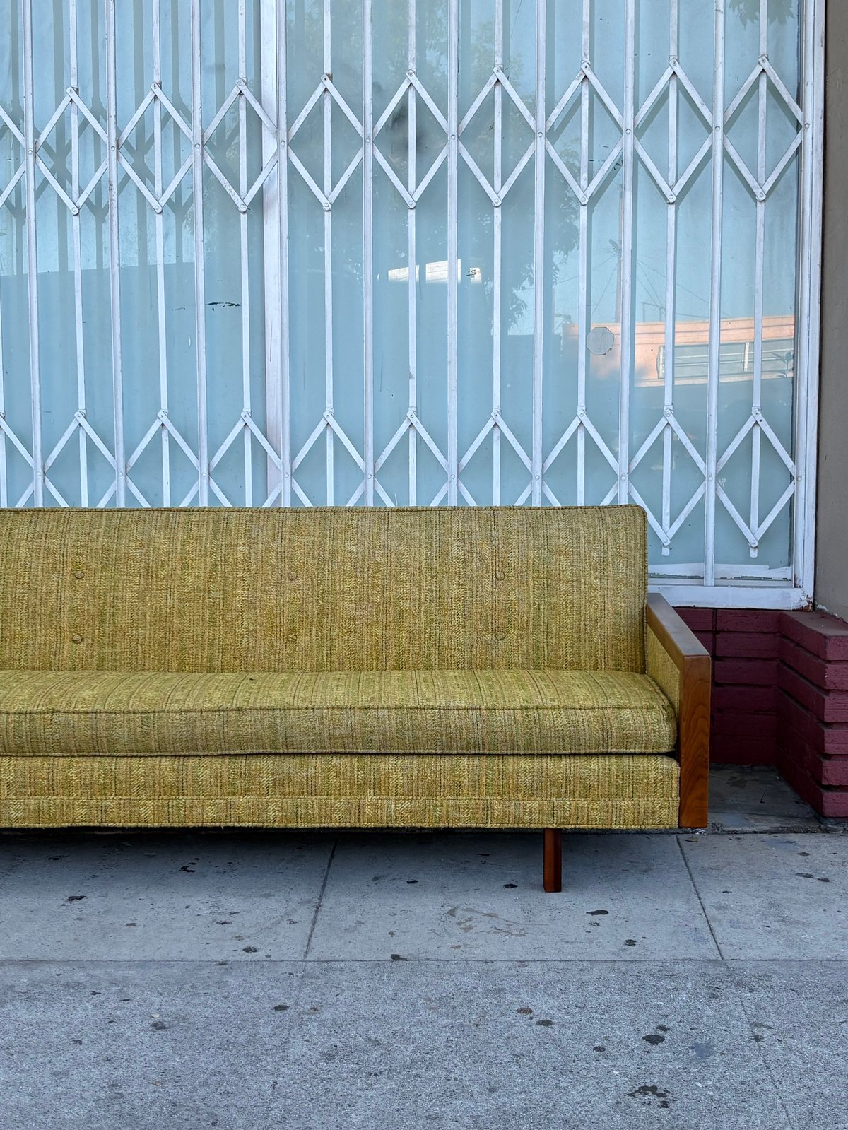 Yellow-green mid-century sofa with wooden frame on a sidewalk, white metal security gate and window behind it