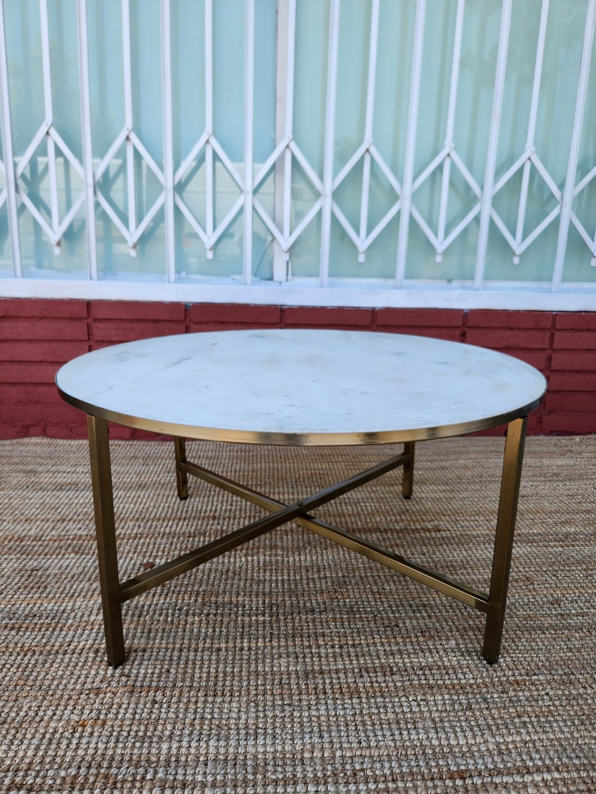 Round marble-top coffee table with brass base and X-frame design on a beige carpet against a brick wall and patterned window