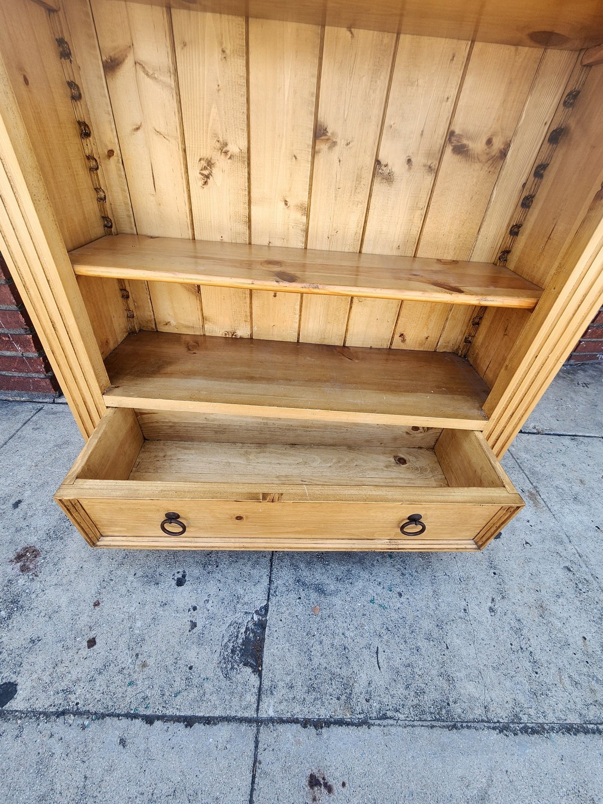 Wooden cabinet with two shelves and a bottom drawer with metal handles, displayed outdoors on concrete.