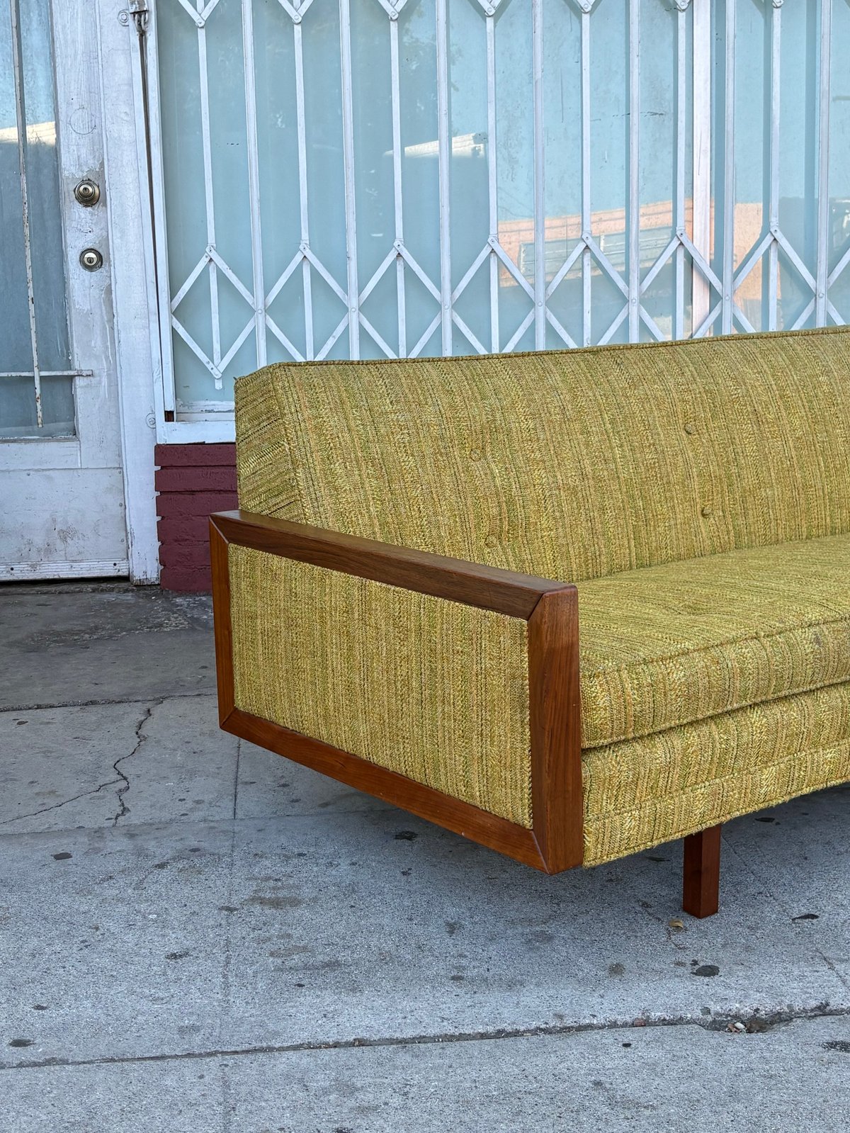 Mid-century modern sofa with yellow-green upholstery and wooden frame sitting on a street corner in front of a blue metal gate