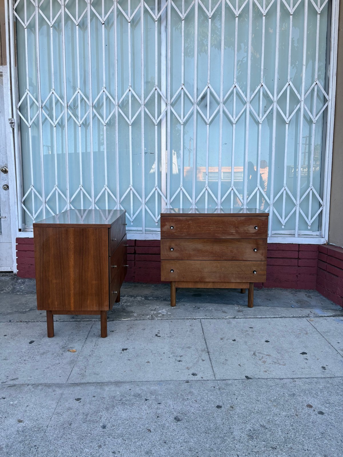 Two mid-century wooden dressers on sidewalk in front of metal security gate storefront
