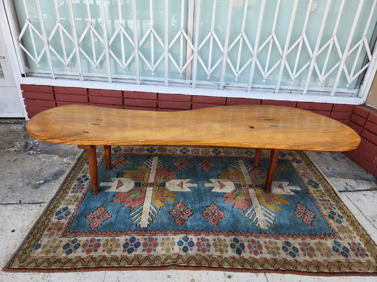 Mid-century wooden surfboard-shaped coffee table on a patterned area rug outdoors against a white gate and red brick wall