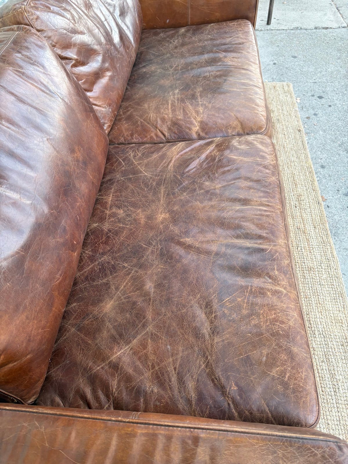Close-up of worn brown leather sofa with cushions showing visible creasing and patina on concrete floor