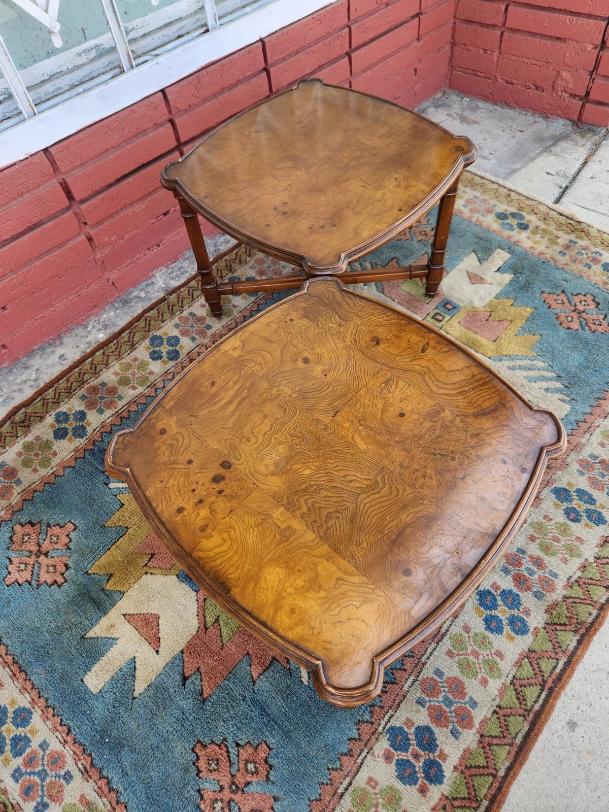 Two vintage wooden tables with scalloped edges on a colorful patterned rug against a red brick wall