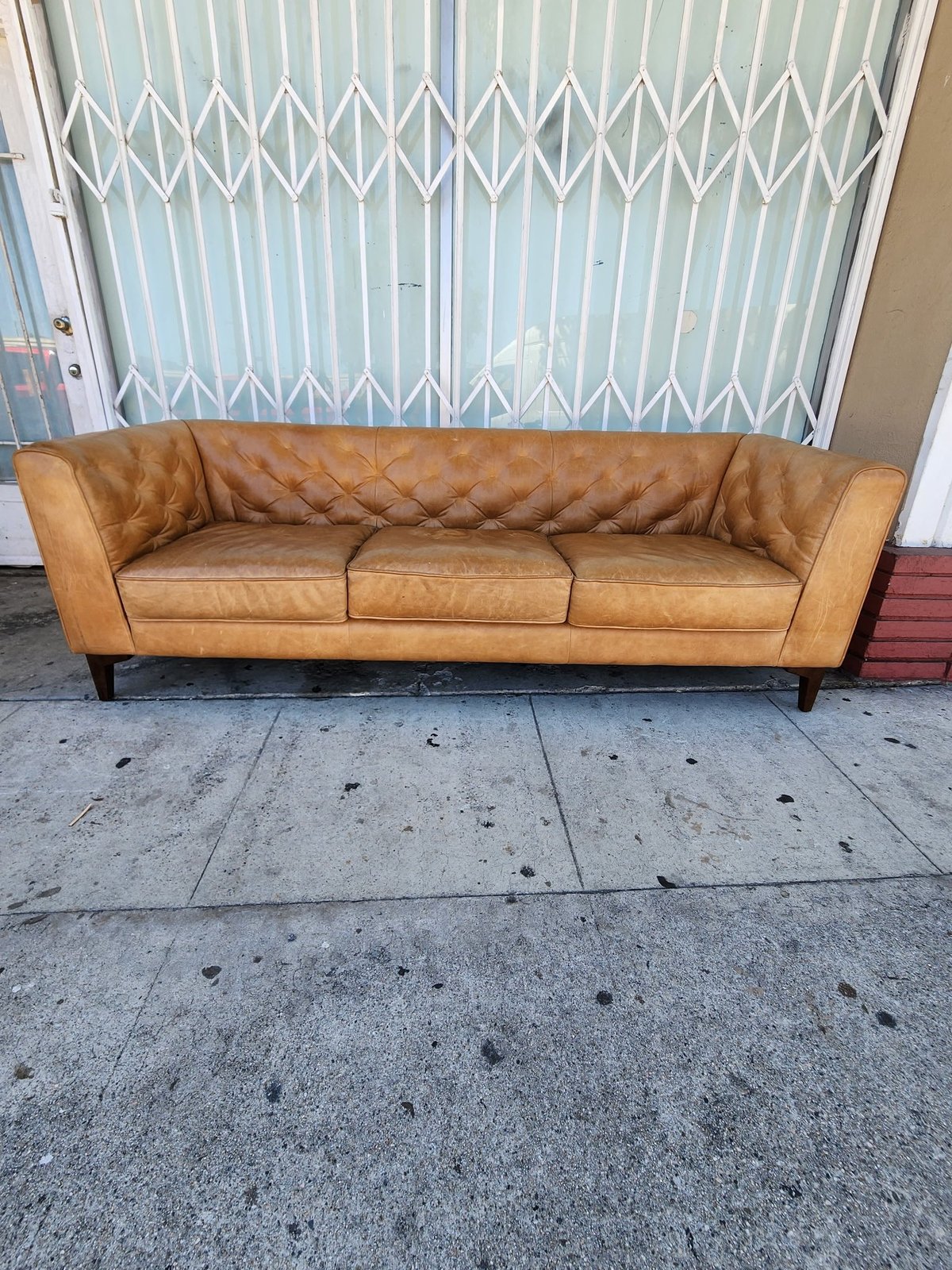 Tan leather quilted sofa with wooden legs sitting on concrete sidewalk in front of white accordion gate