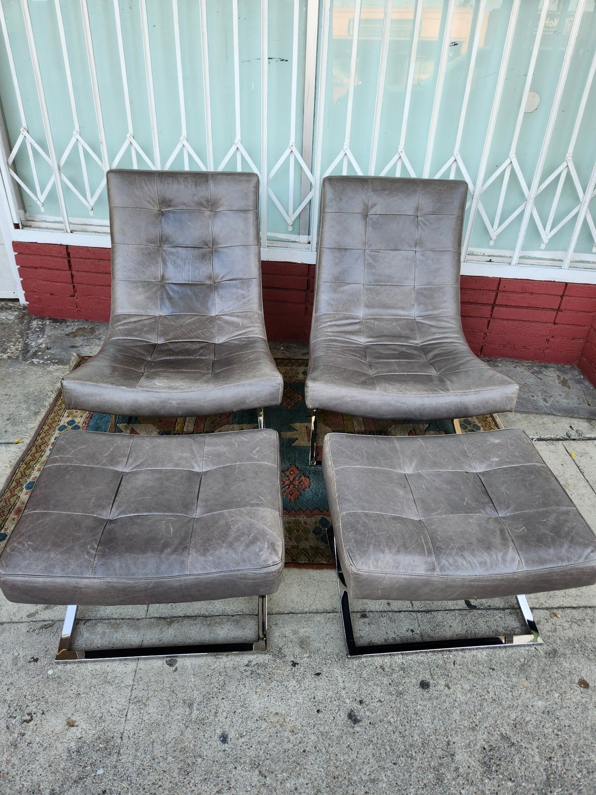 Two pairs of grey upholstered lounge chairs with matching ottomans on concrete pavement against a corrugated metal fence