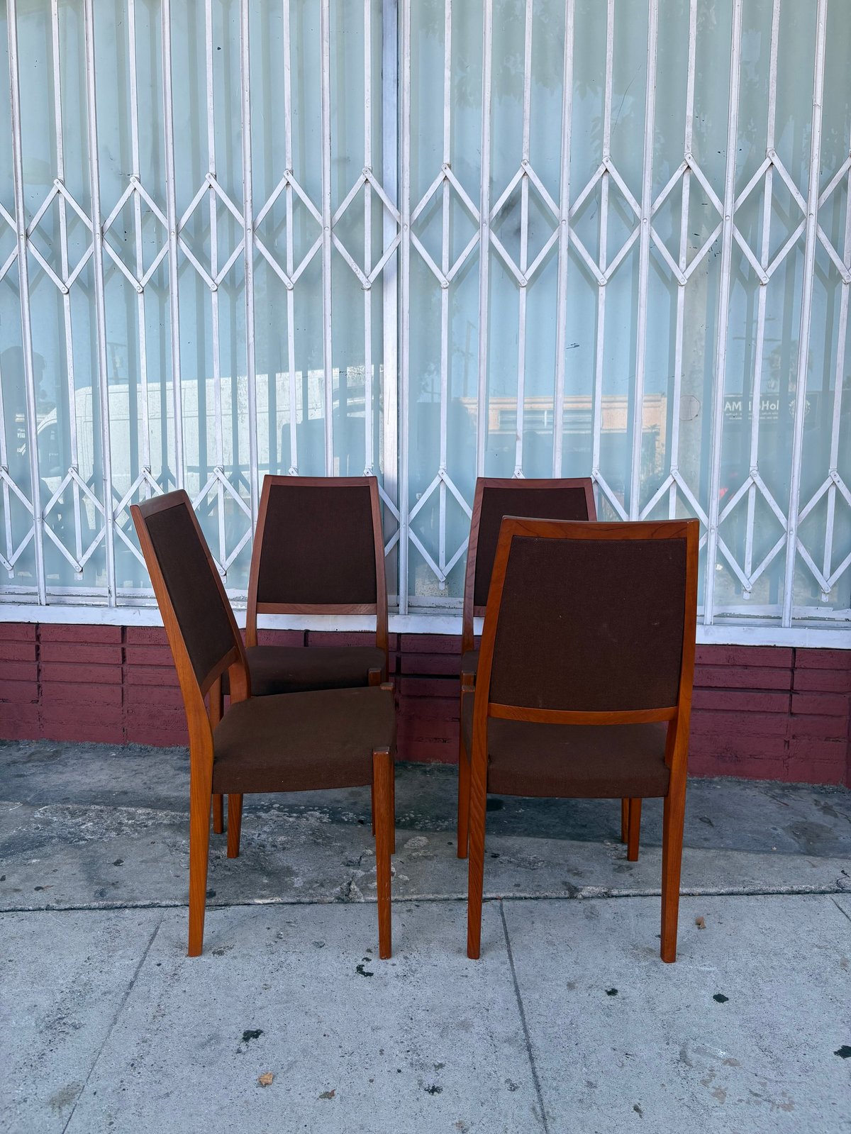 Four wooden chairs with brown upholstered seats on a concrete sidewalk in front of a white metal gate