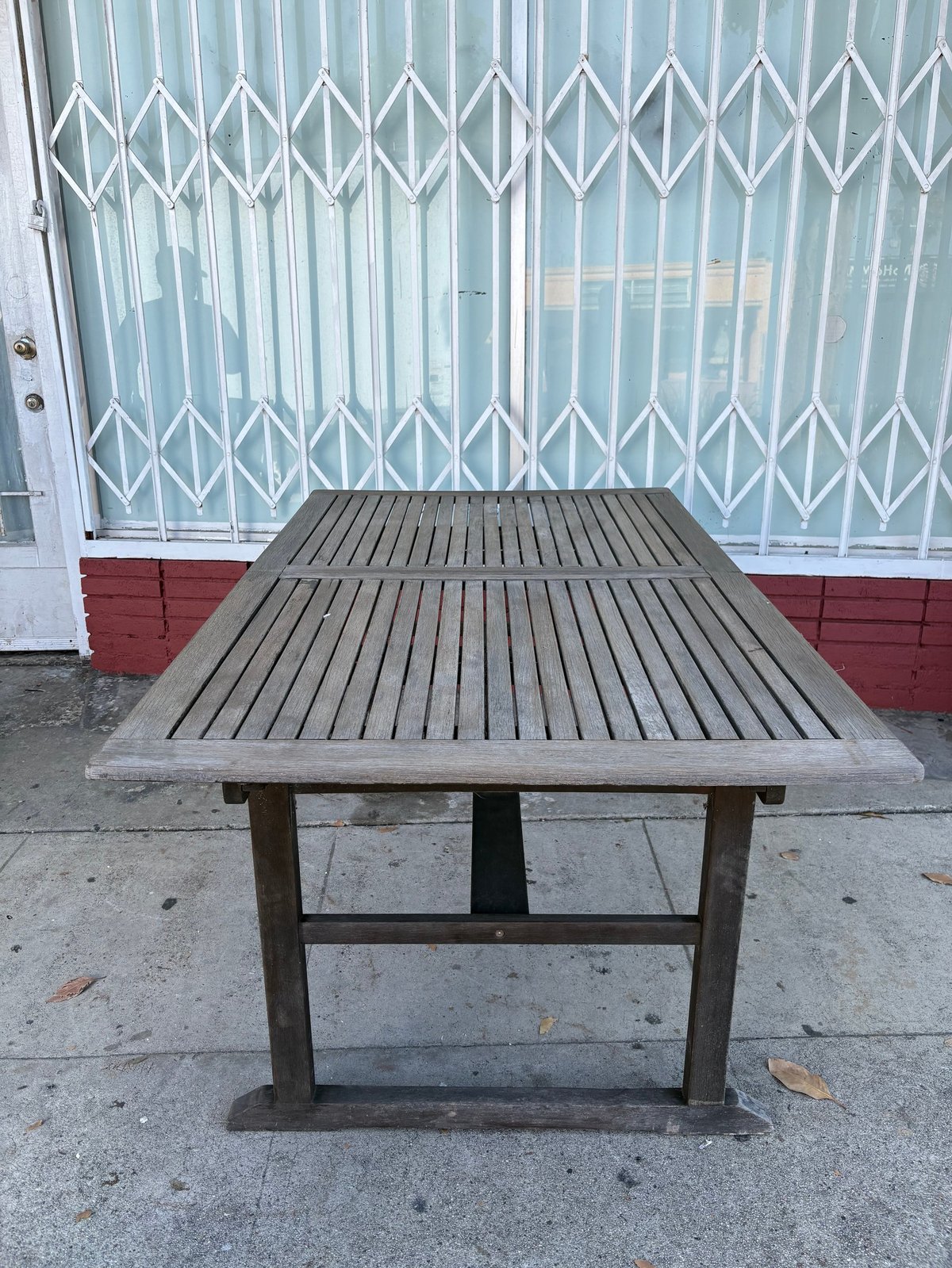 Wooden picnic table on concrete pavement with white metal security gate and red wall in background