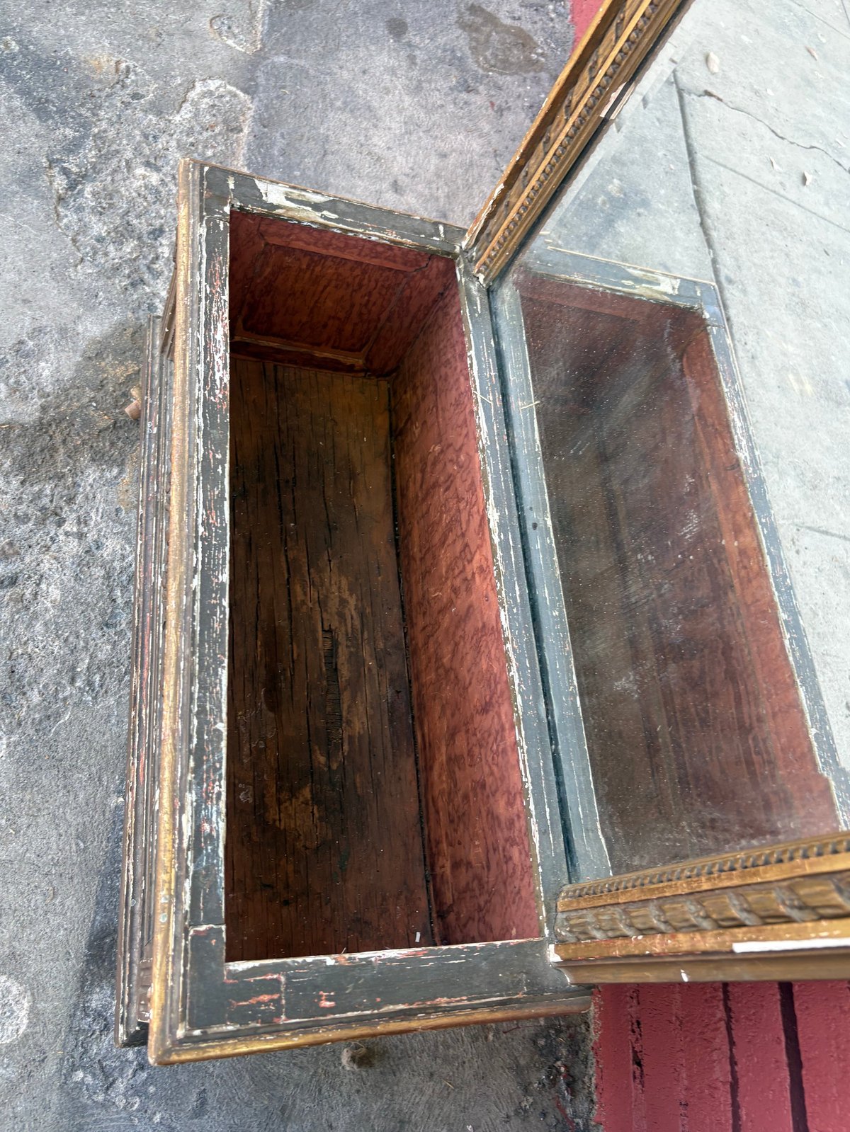Empty wooden box with burgundy velvet lining and ornate gold frame, open on concrete surface.