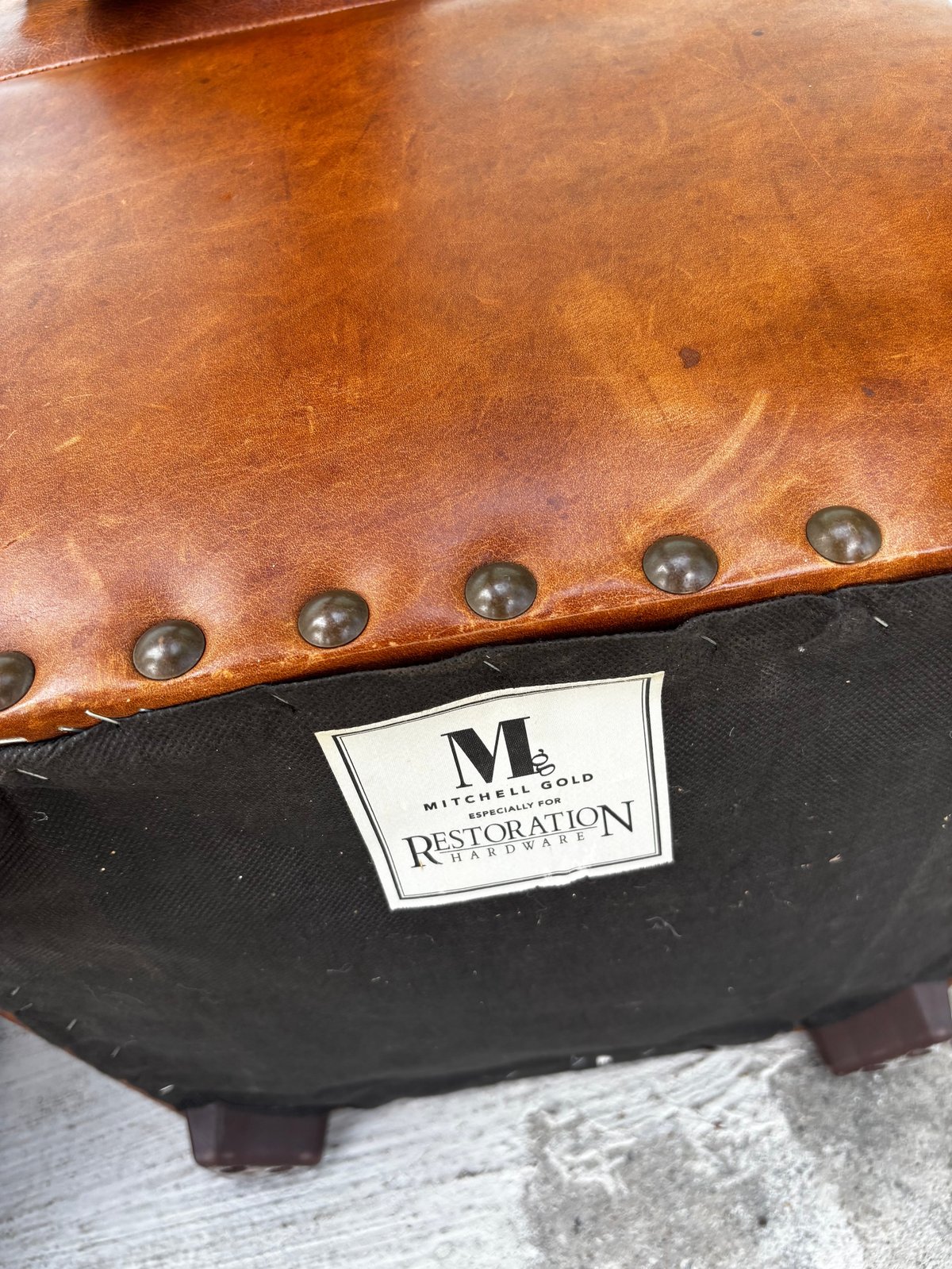 Close-up of a tan leather furniture piece with studded trim and a Mitchell Gold Restoration label