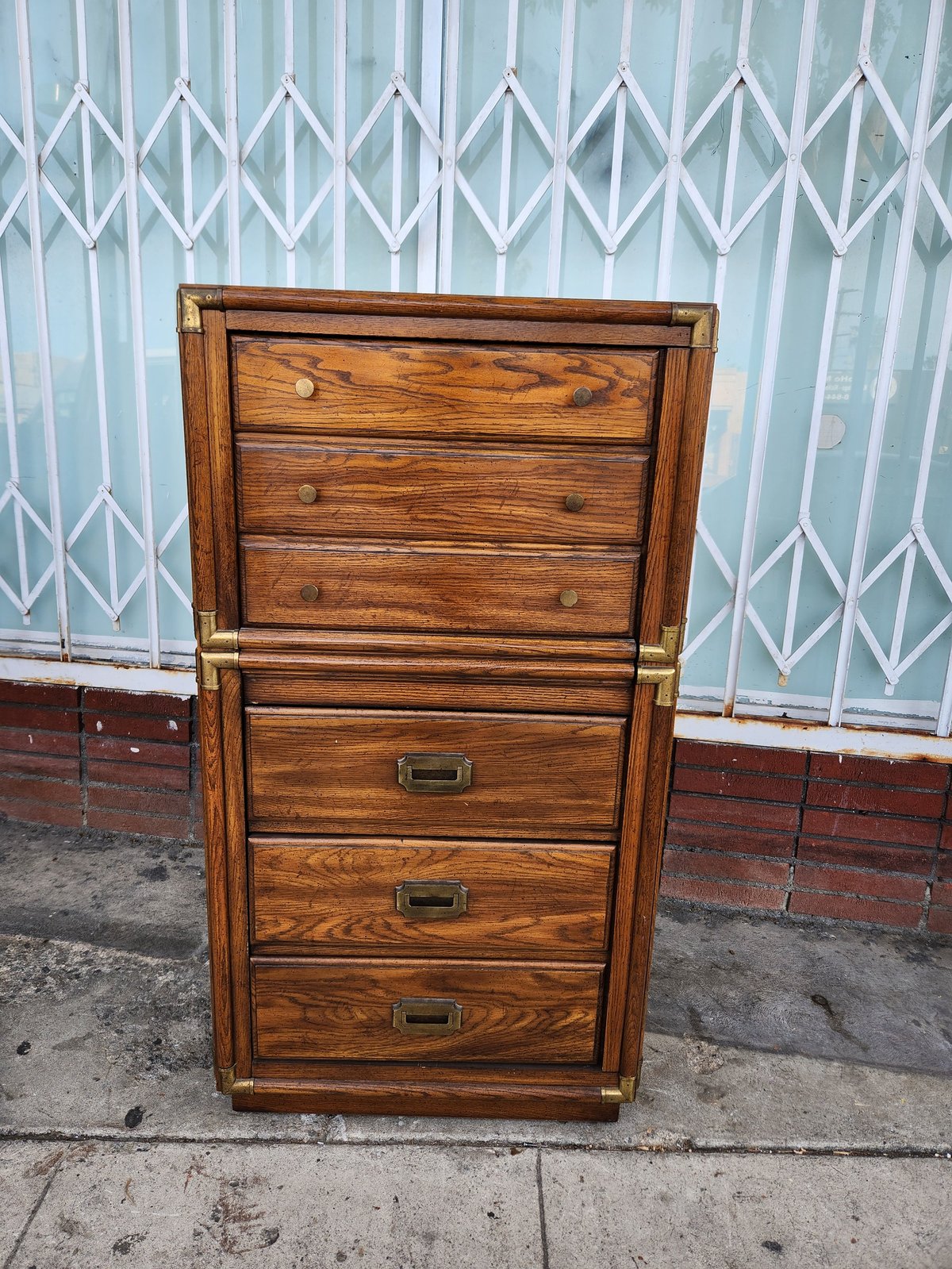 Wooden dresser with six drawers and brass handles against a weathered metal door and brick wall