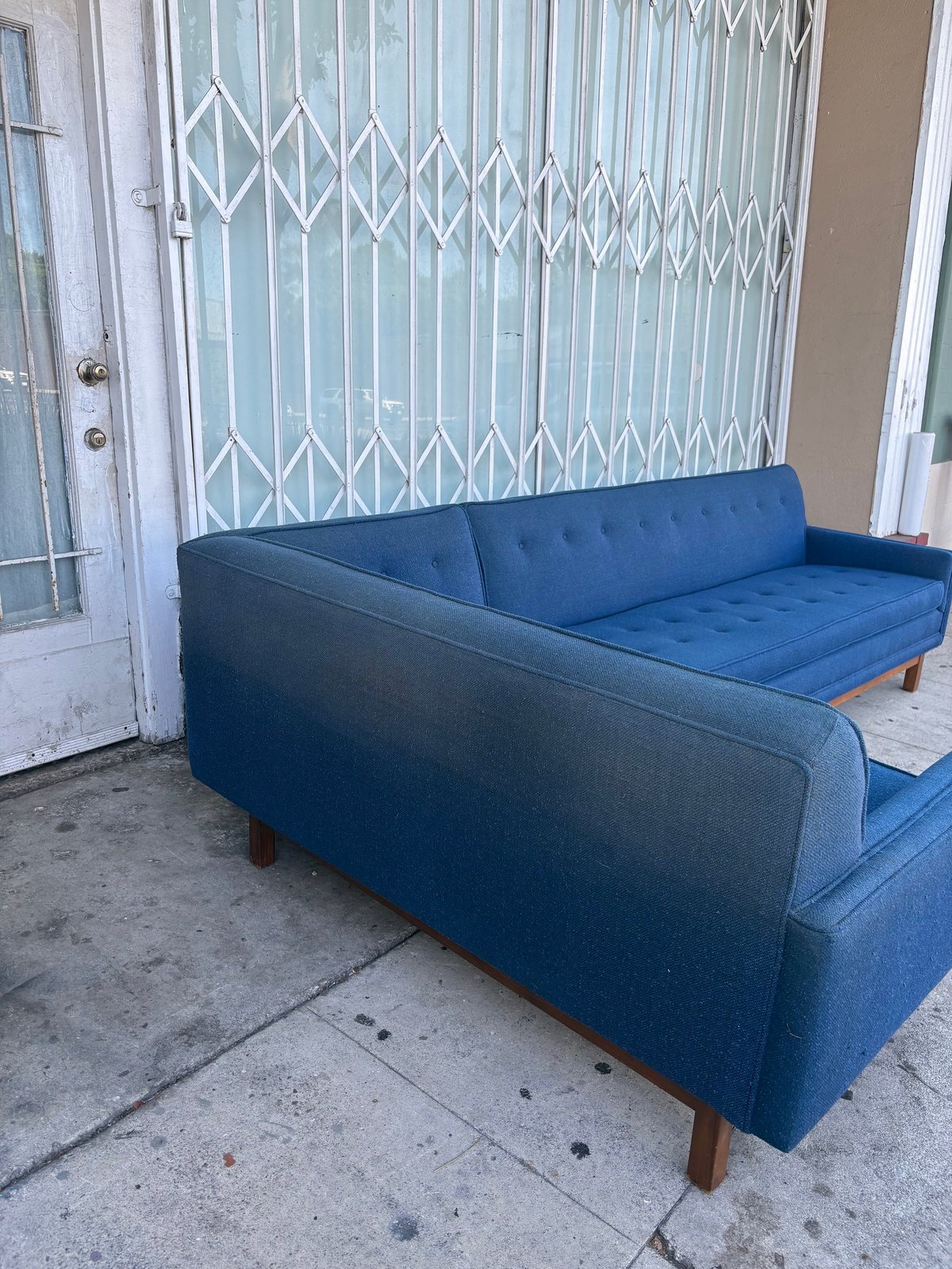 Blue mid-century modern sofa with wooden legs on outdoor concrete patio, white metal security door and gate in background