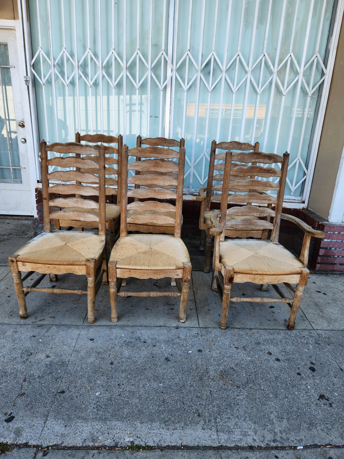 Three wooden ladder-back chairs with woven rush seats lined up outdoors against a light blue metal gate