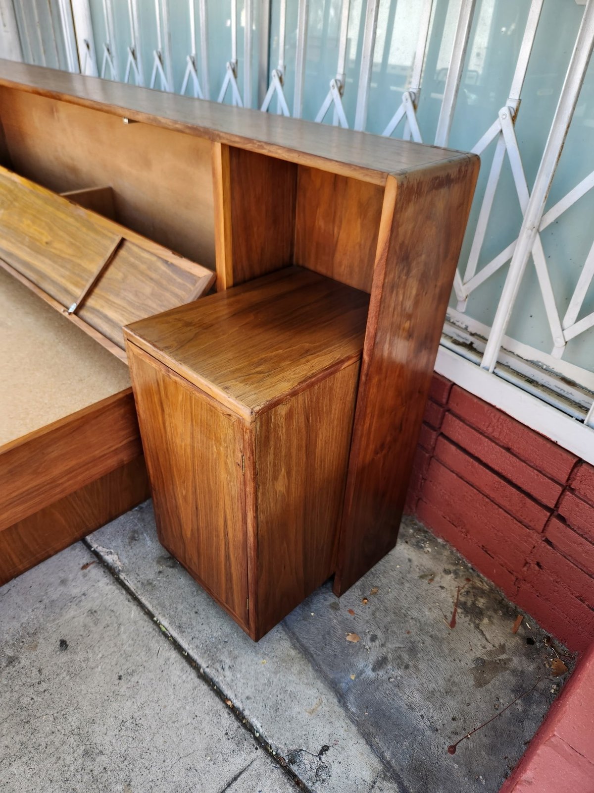Vintage wooden shelf unit with open compartments and cabinet doors, displayed outdoors next to a blue and white metal gate