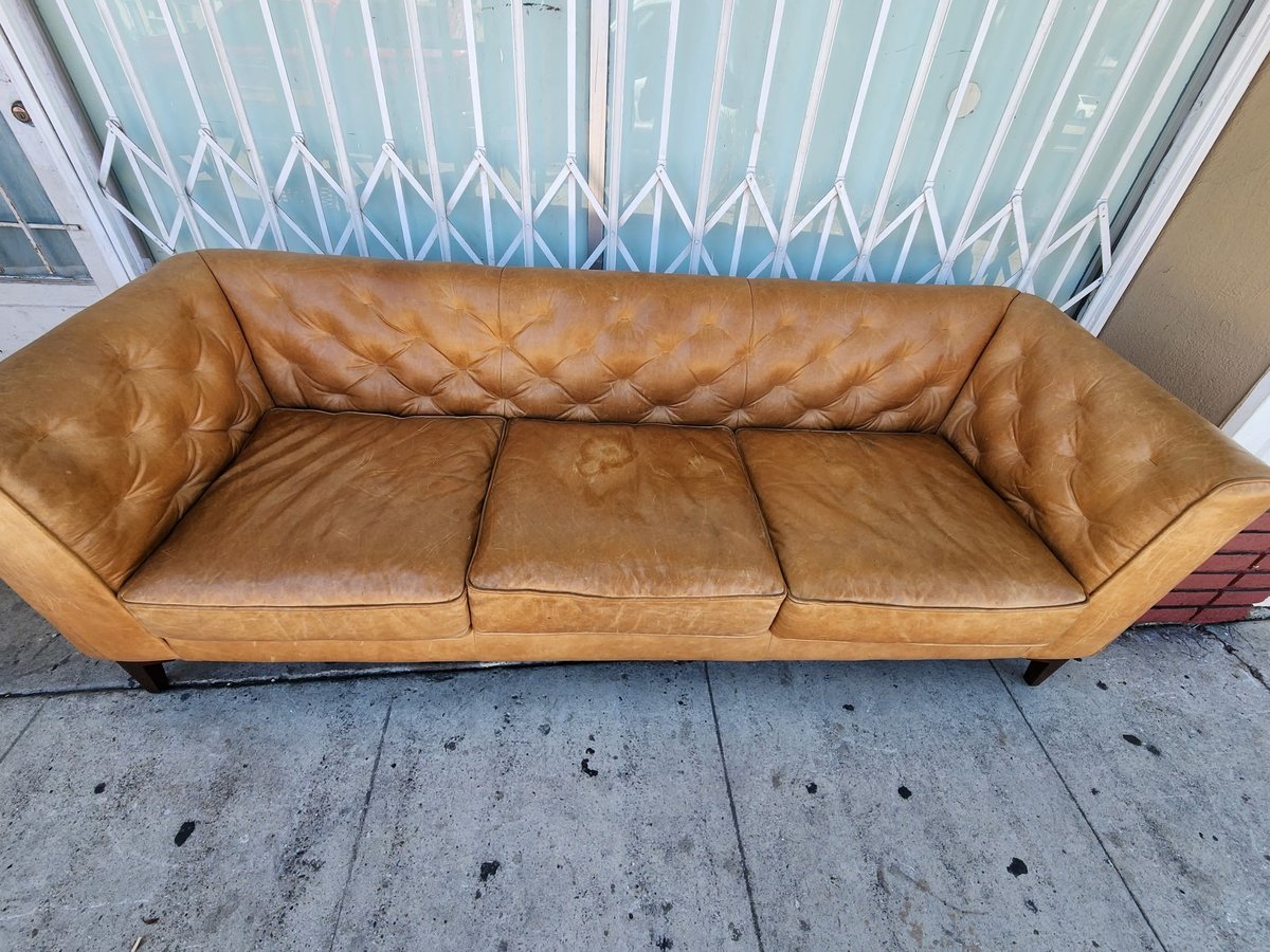 Tan leather three-seater sofa with quilted back and cushioned seats on concrete floor against striped window blinds
