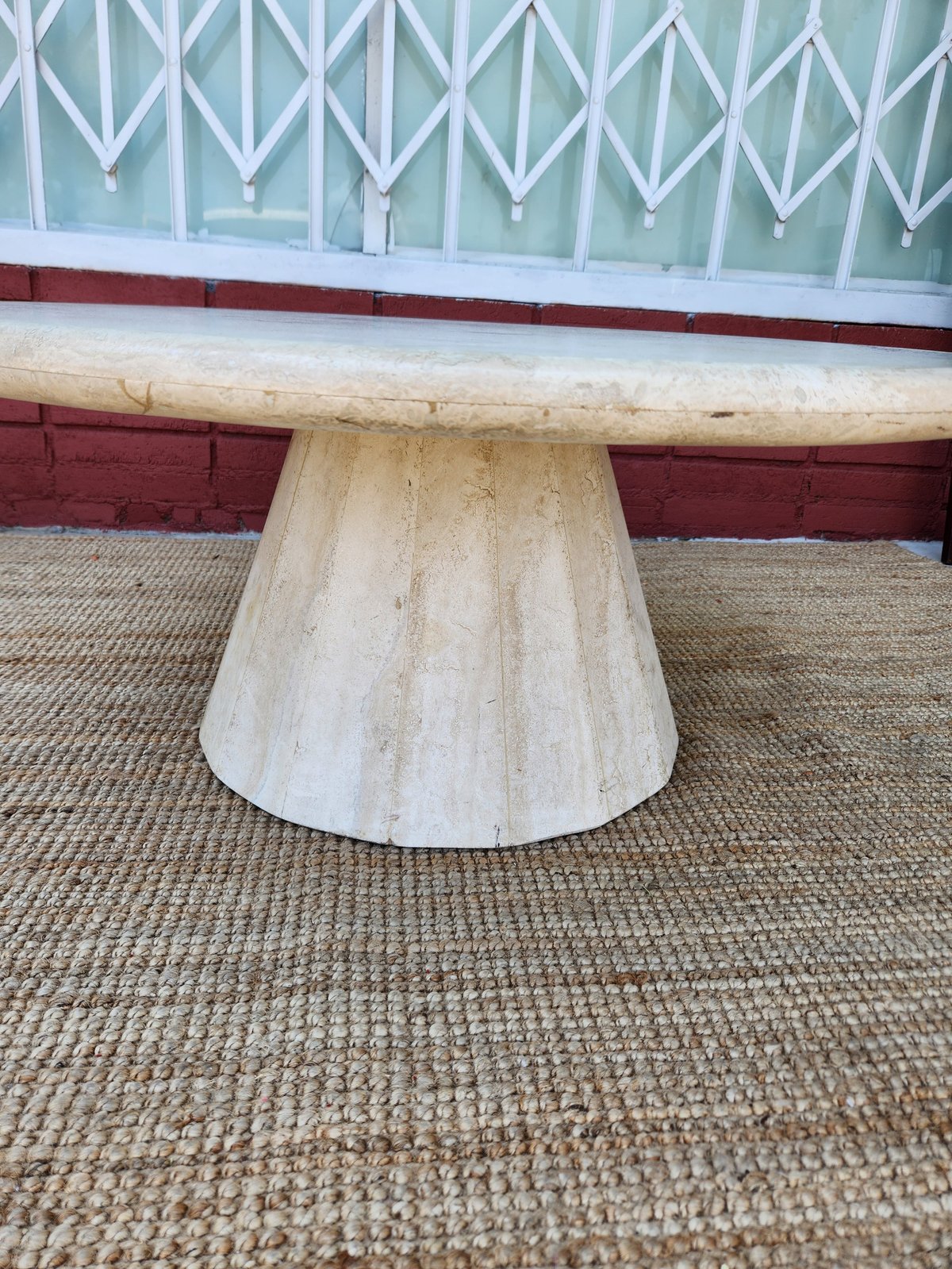 Travertine cone-shaped pedestal table base on beige carpet with red bench seating and patterned windows in background