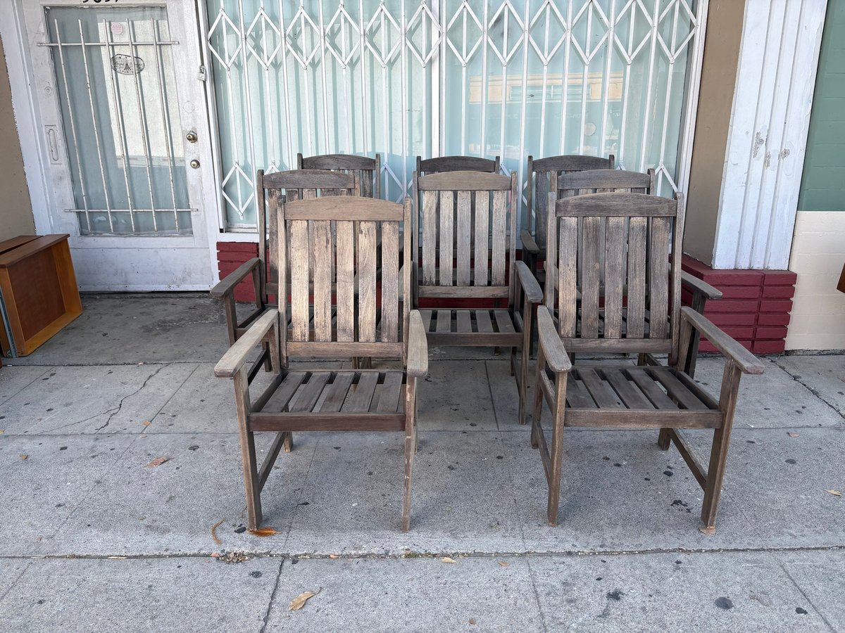 Five wooden chairs arranged on a sidewalk in front of a storefront with metal security gates and glass doors
