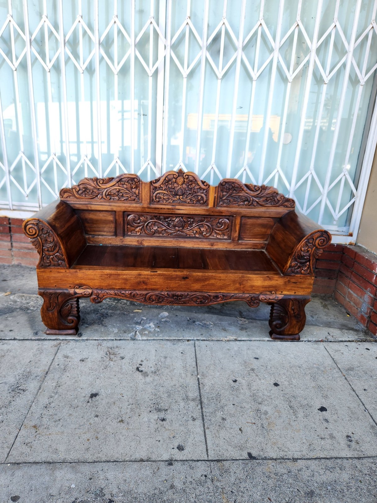 Ornately carved wooden bench with intricate details, positioned against white accordion curtains on a concrete patio