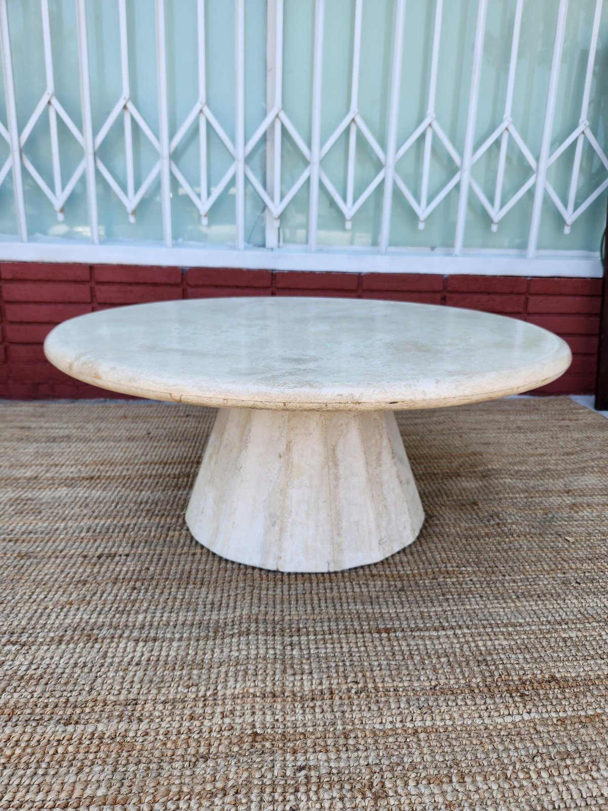 Whitewashed wooden round pedestal dining table on a beige rug against a red brick wall and metal lattice gate