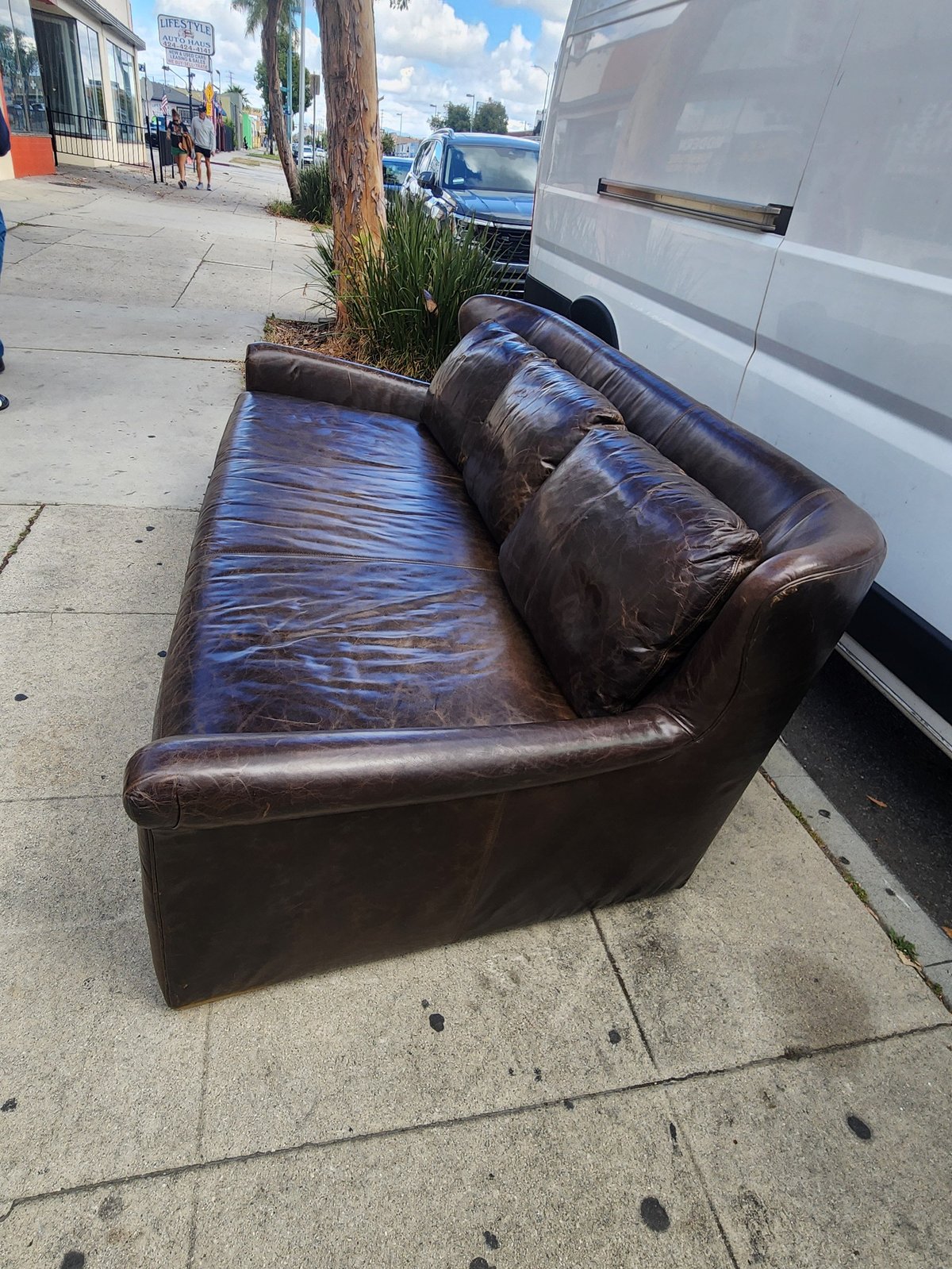 Worn brown leather couch sitting on a sidewalk next to a white van and parked cars