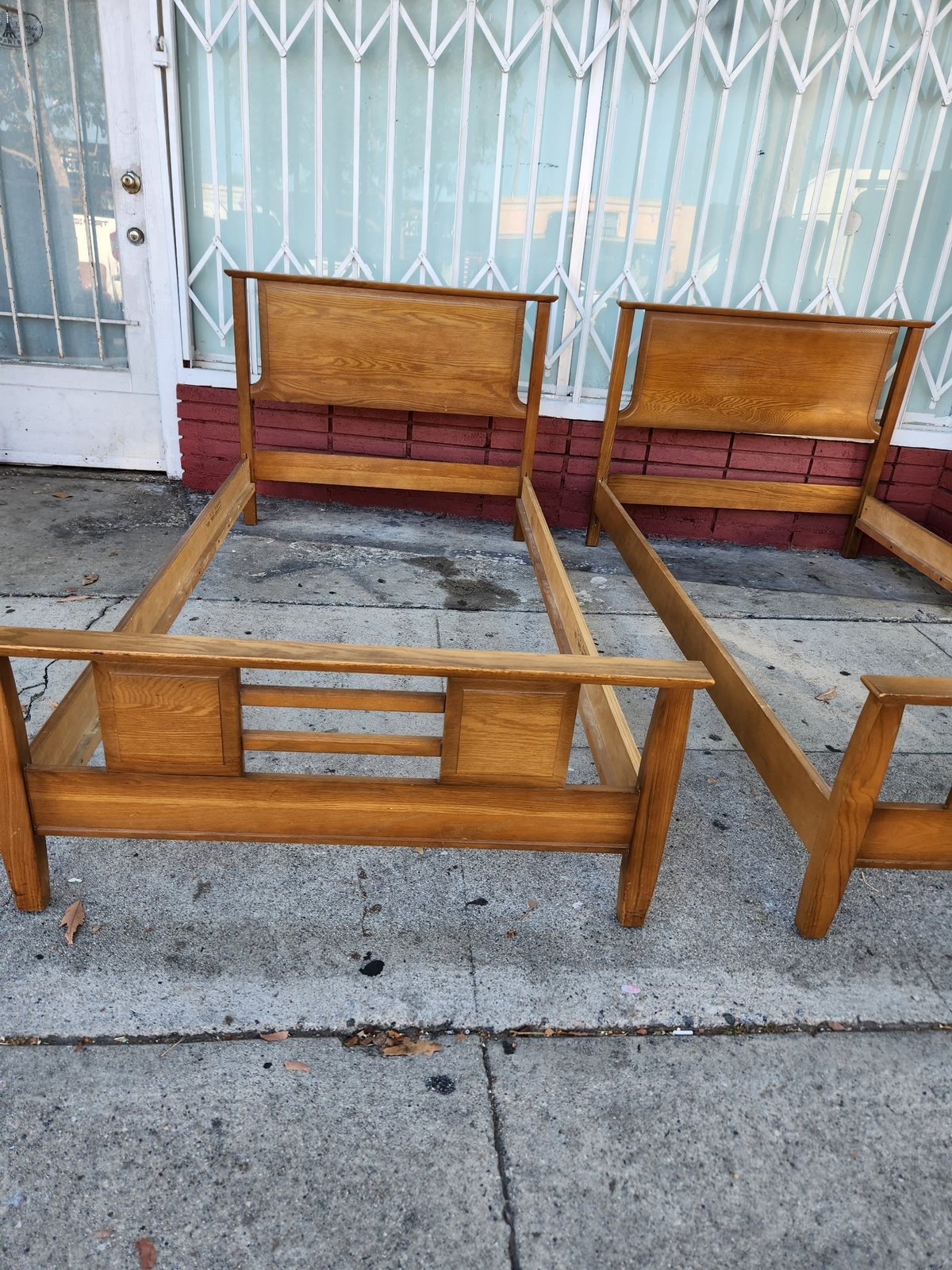 Two wooden bed frames with red upholstered panels displayed on a sidewalk outside a storefront with metal grating doors