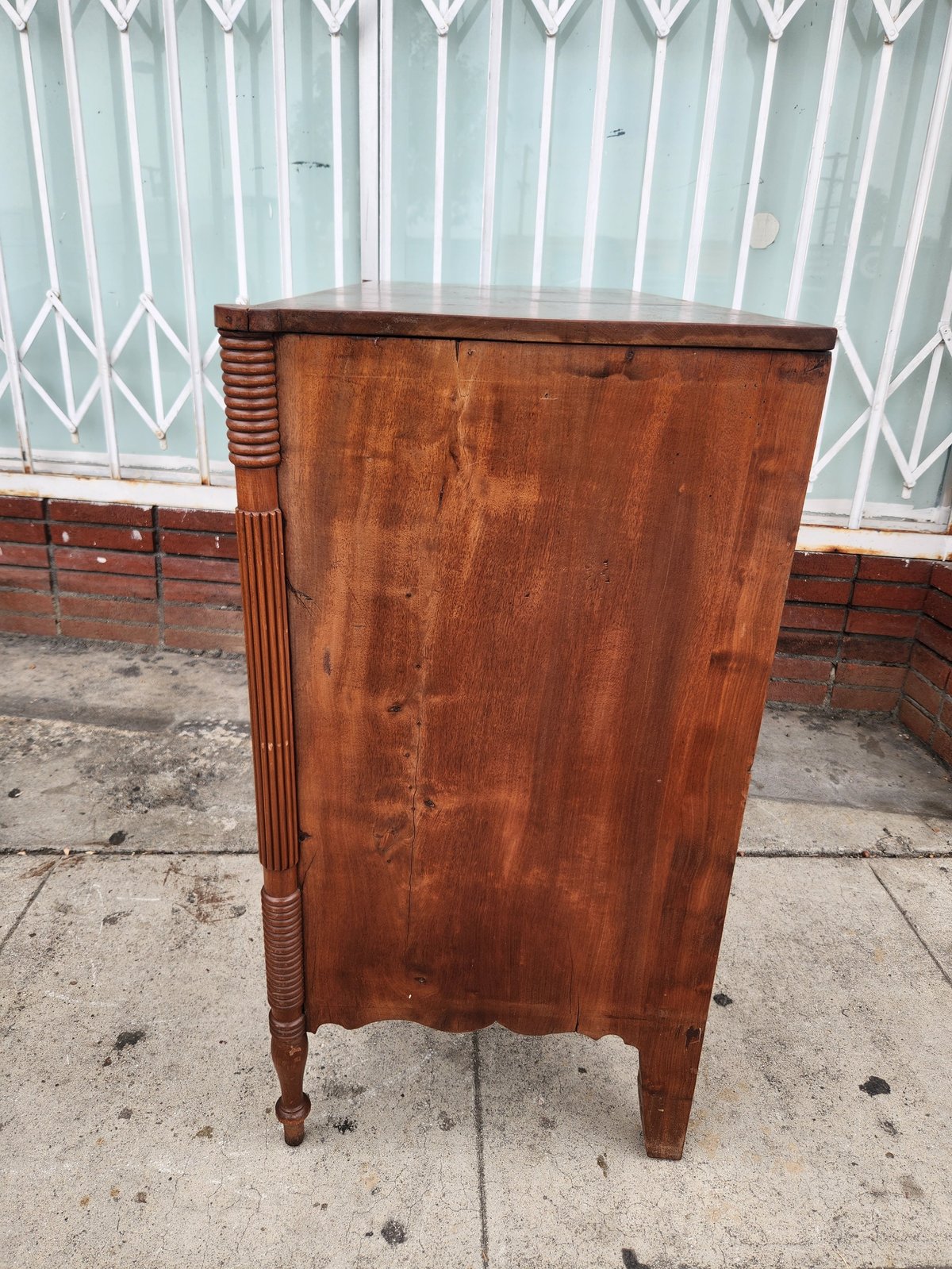 Antique wooden cabinet with carved leg details on concrete driveway with metal gate in background
