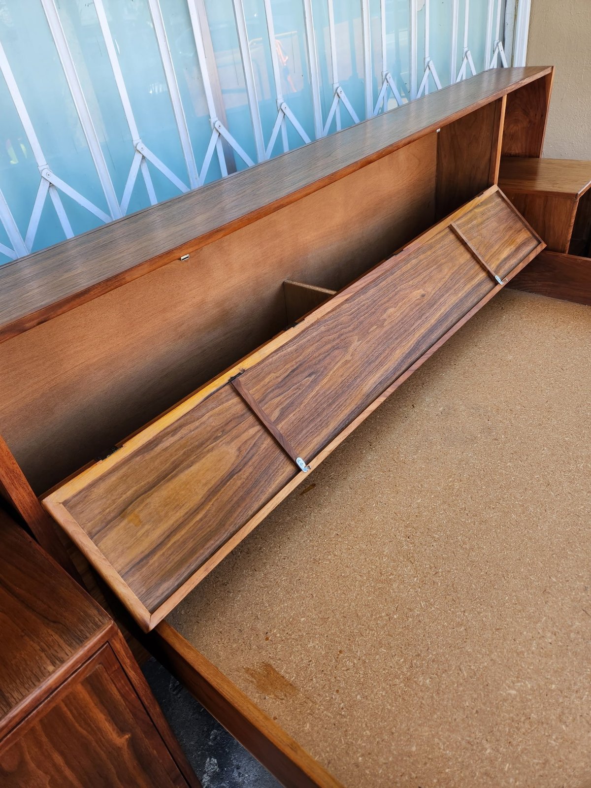 Wooden storage bench with hinged lids open, revealing compartments inside, positioned under a window with blue striped curtains