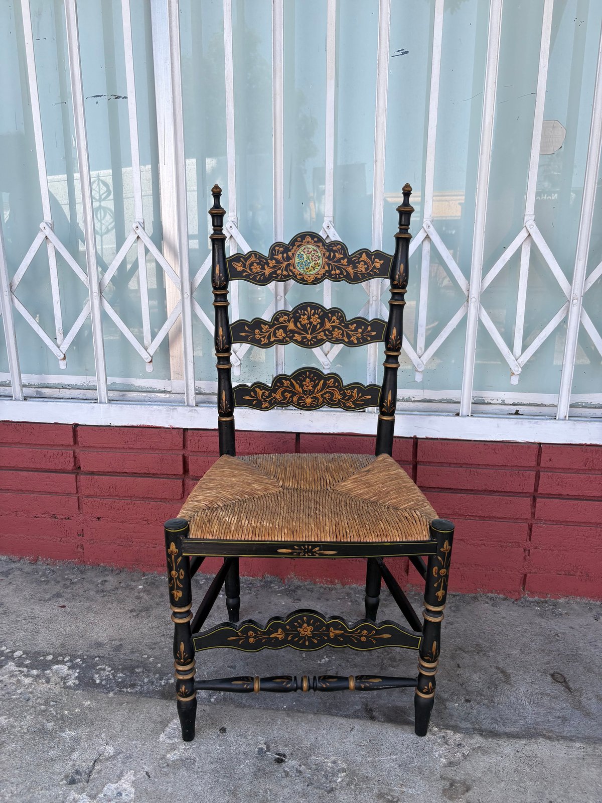 Ornate black wooden chair with woven rush seat against a weathered storefront with white lattice and red brick