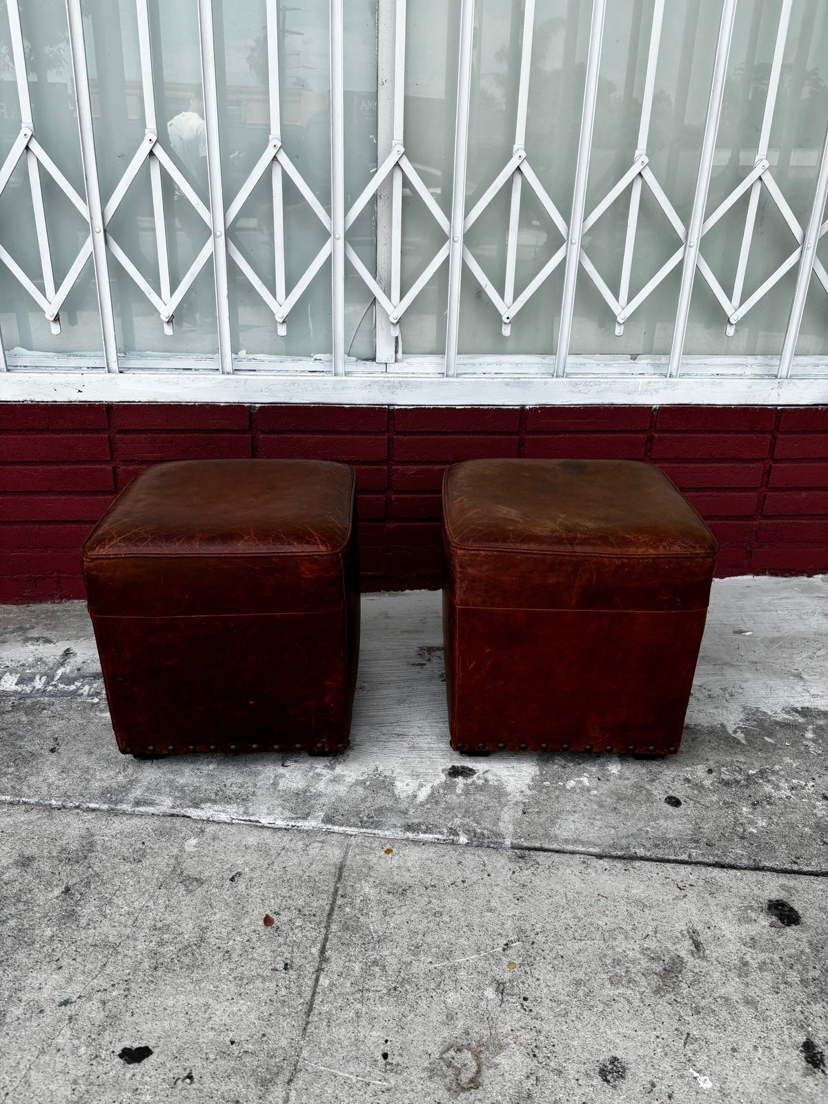 Two worn burgundy leather ottomans positioned side by side on concrete, with a white lattice door and red brick wall behind them