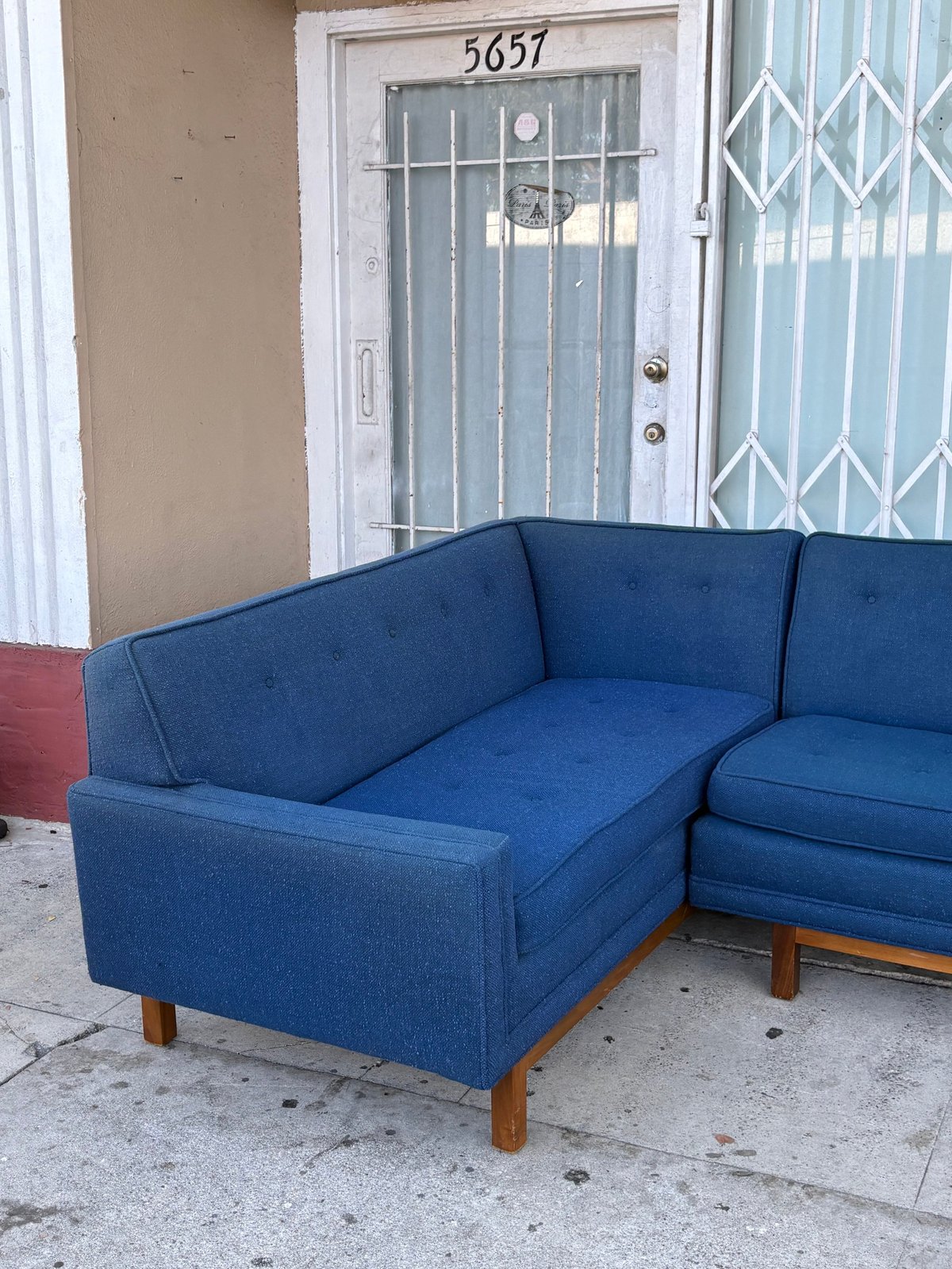 Blue upholstered sectional sofa with wooden legs on a concrete porch outside a white door