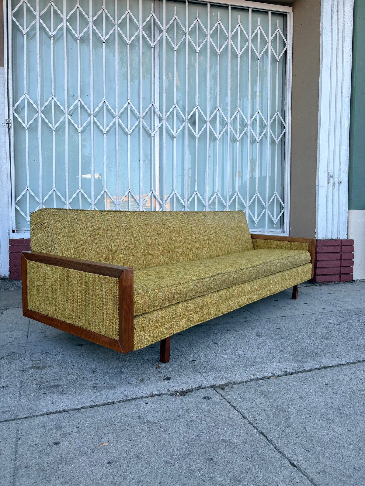 Vintage mid-century yellow-green upholstered sofa with wooden arms on a sidewalk in front of a metal security gate