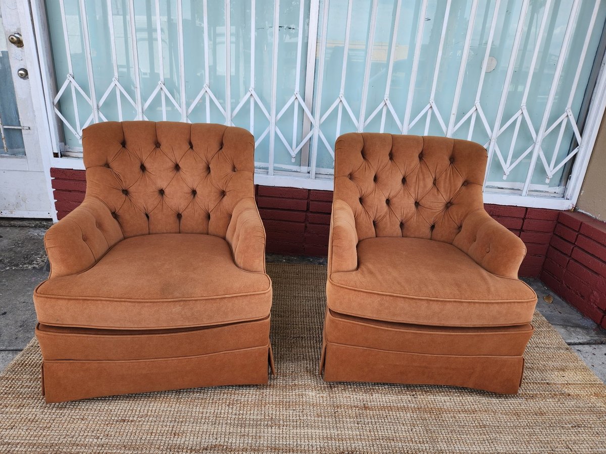 Pair of orange-brown tufted armchairs with rolled arms on a beige rug, with white shuttered windows and red wall behind