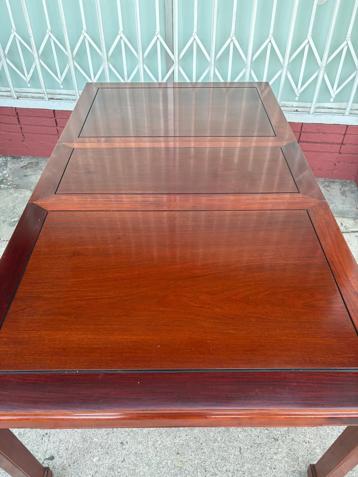 Wooden dining table with glass top sections and removable leaves against a metal gate and red wall