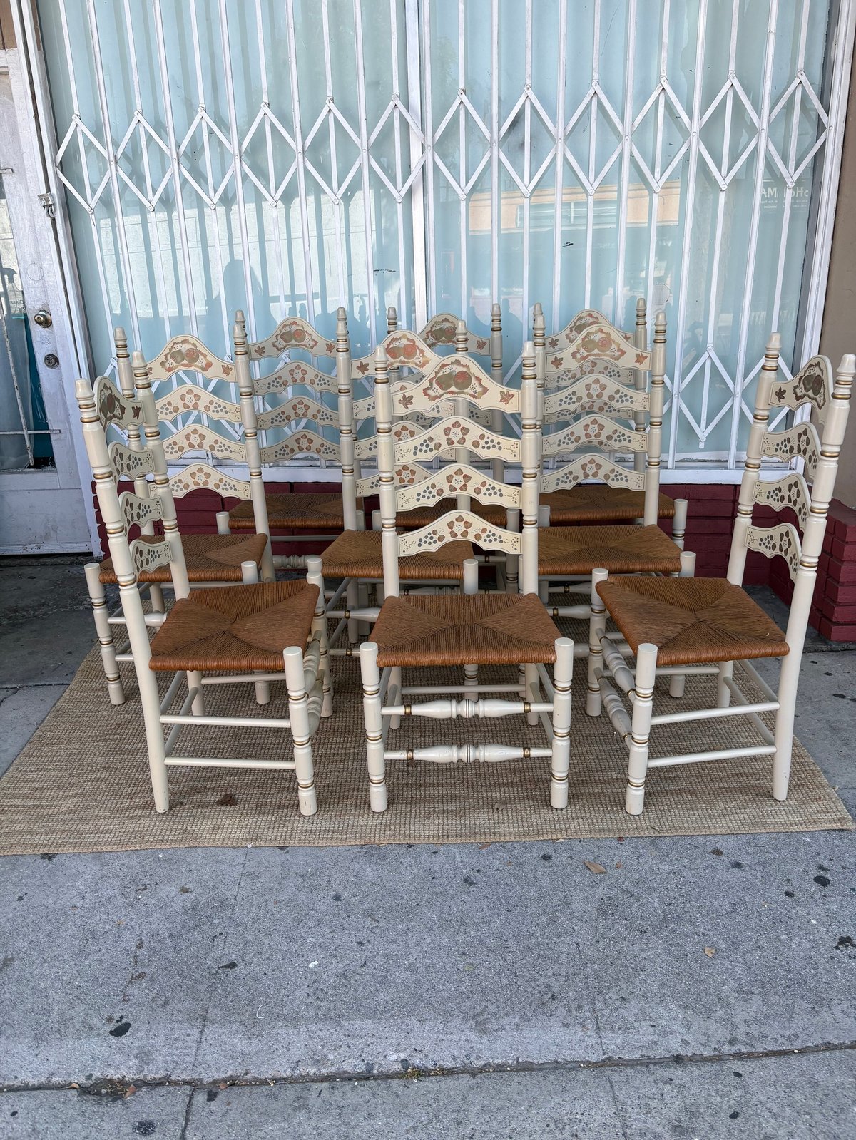 Set of six wooden chairs with white frames, woven rush seats, and carved decorative backs displayed on a doormat outdoors.