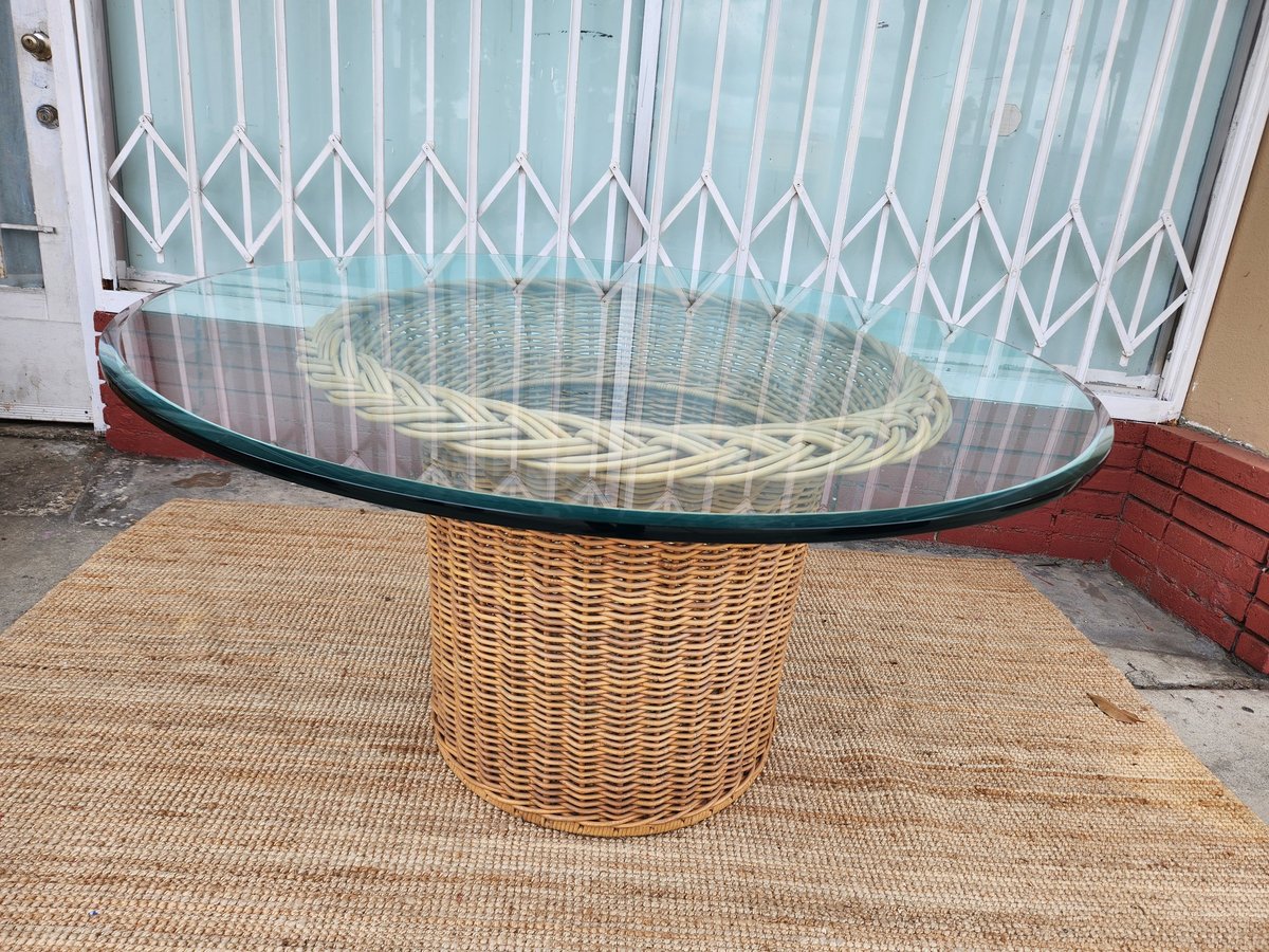 Round glass-top dining table with woven wicker base on outdoor patio near white gate