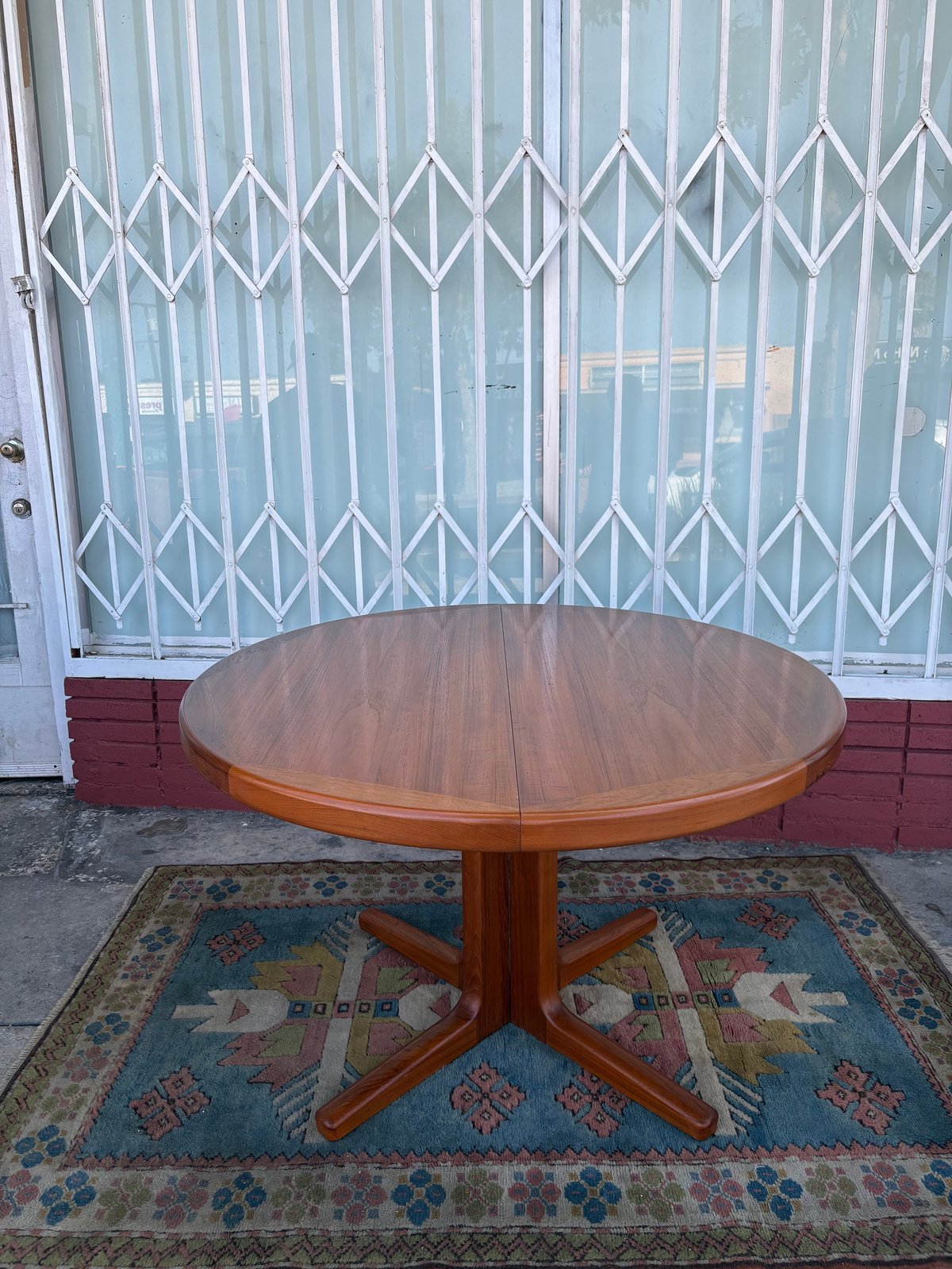 Round wooden dining table with pedestal base on colorful rug, white metal gate behind