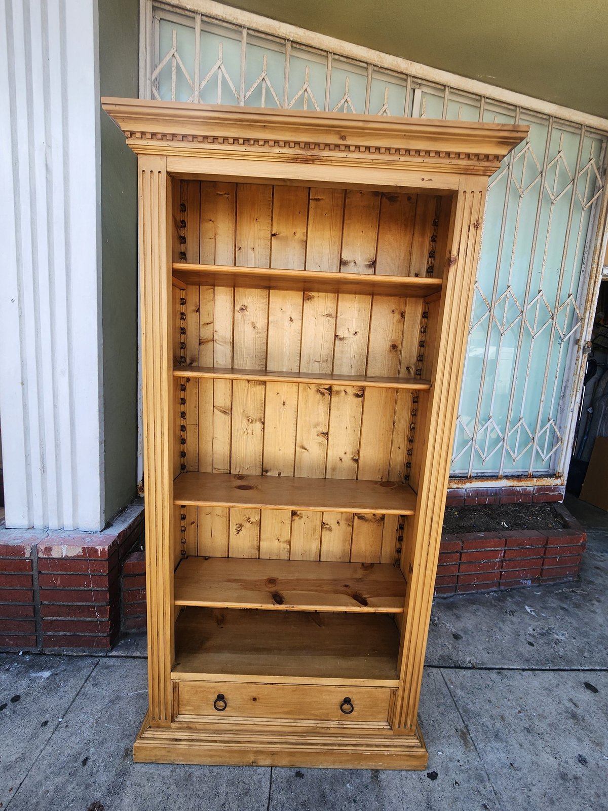Wooden bookshelf with decorative crown molding, adjustable shelves, and lower storage drawer displayed outdoors