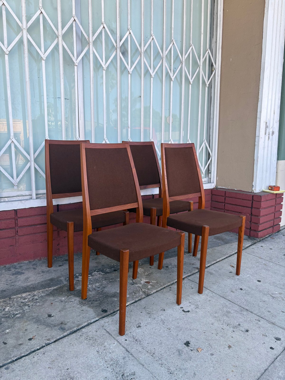 Four wooden dining chairs with brown upholstered seats arranged on a sidewalk in front of a storefront with metal security gates