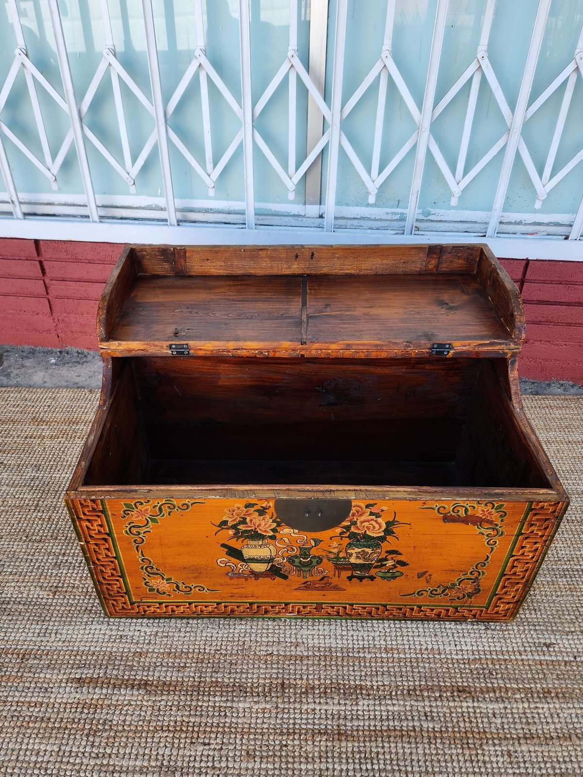 Antique wooden trunk with decorative Asian-inspired artwork on front, open lid revealing empty storage space, against red brick wall