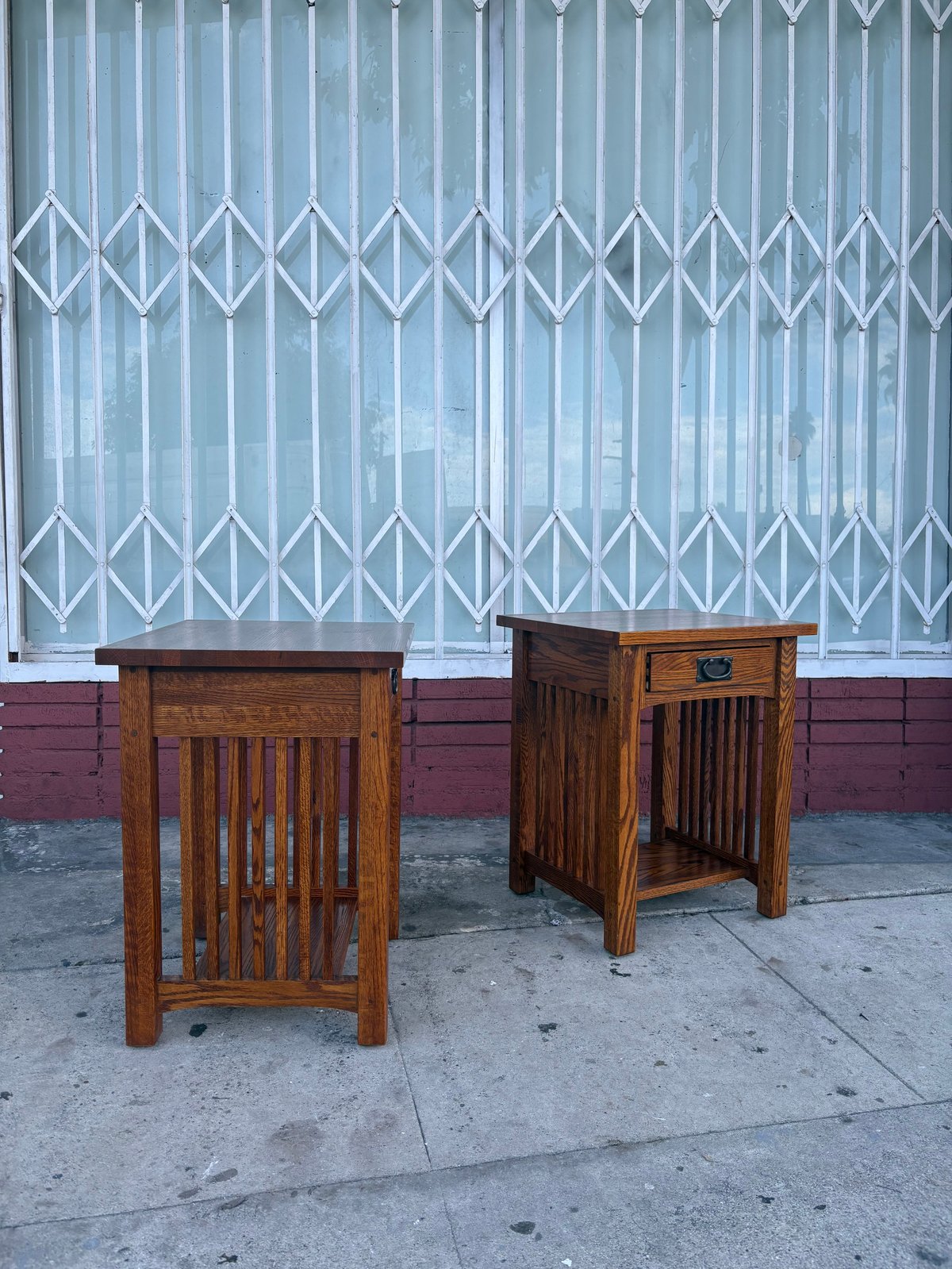 Two wooden side tables with vertical slat details displayed on a sidewalk in front of a metal corrugated security door with maroon lower panels