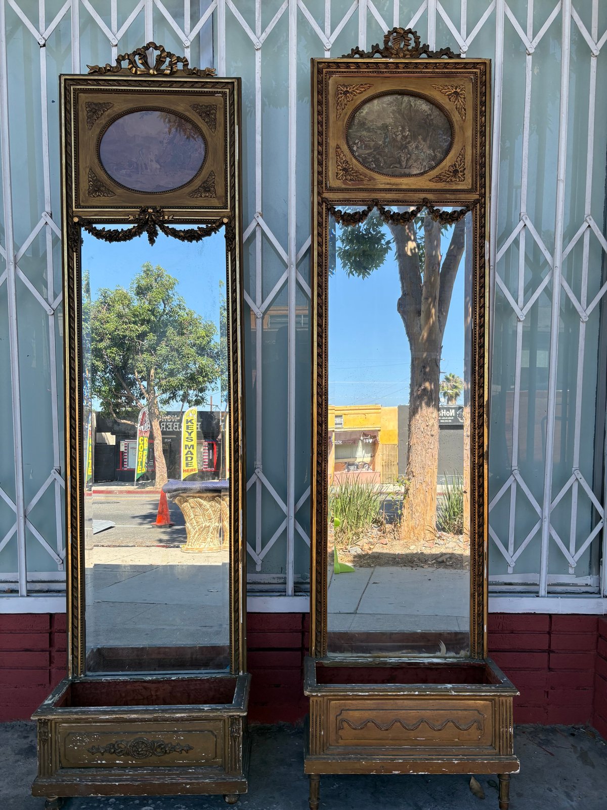 Two ornate vintage mirrors with oval tops and decorative frames reflecting outdoor buildings and trees against a geometric patterned wall