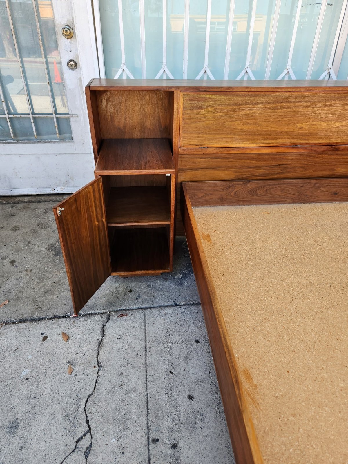 Wooden bedroom set with open cabinet door showing interior shelves, positioned on a concrete driveway with white garage door visible
