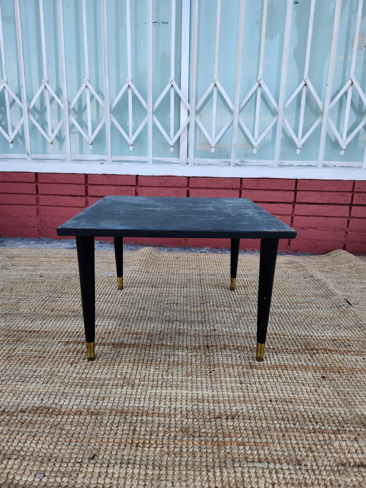 Black rectangular vintage table with tapered legs and brass feet on concrete ground against red brick wall and white geometric gate