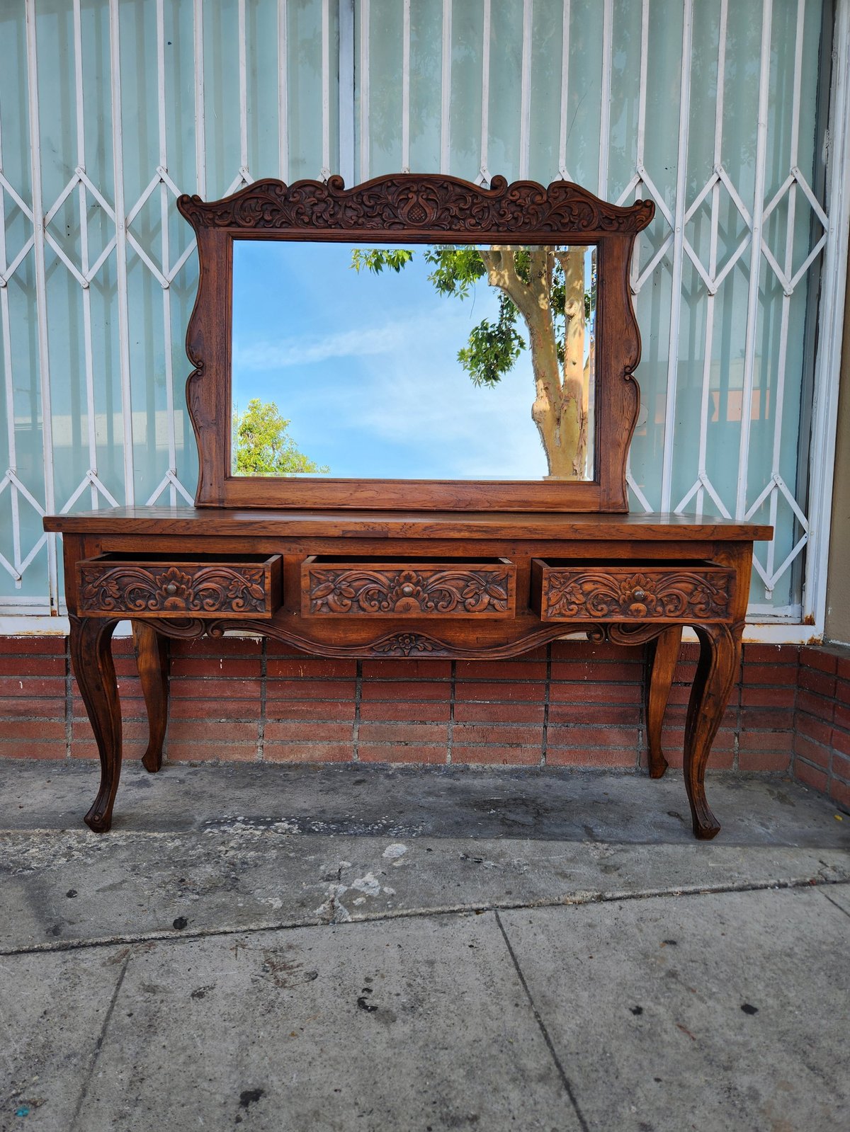Ornate wooden vanity with decorative mirror and carved drawers displayed on a sidewalk against a metal gate
