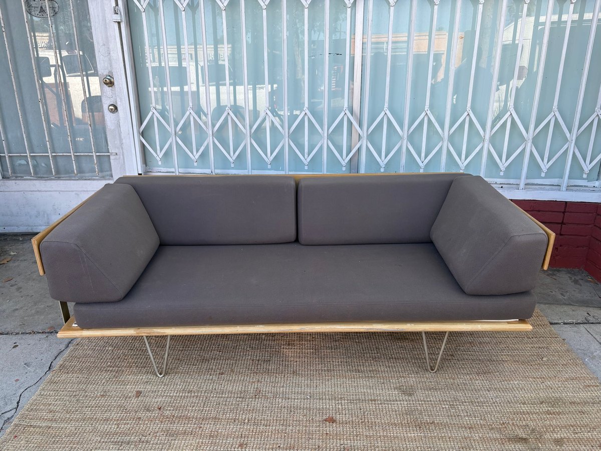 Gray mid-century modern sofa with wooden frame and metal legs on a tan rug, displayed on a driveway with metal gate in background