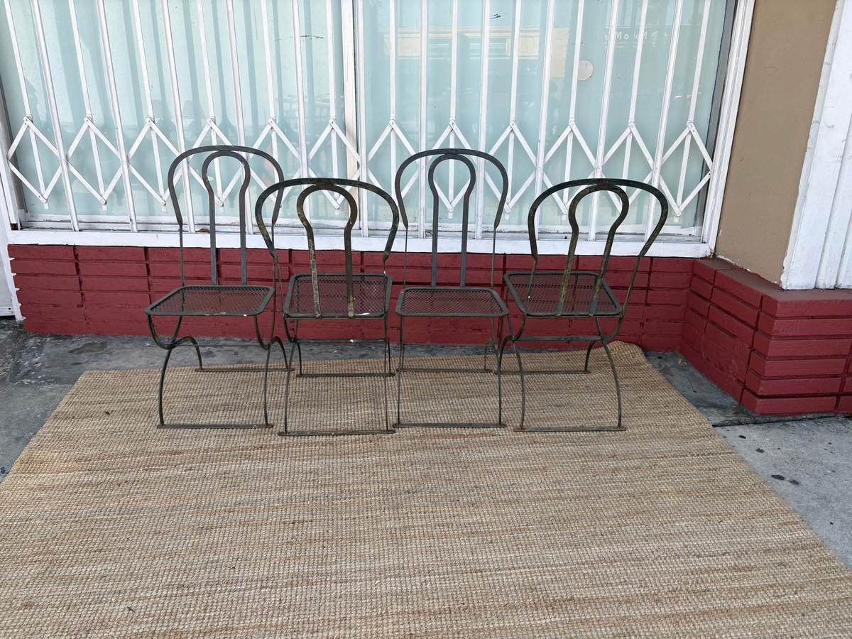 Four metal outdoor chairs with curved backs arranged on a beige outdoor mat in front of a building with red wall trim and louvered window blinds