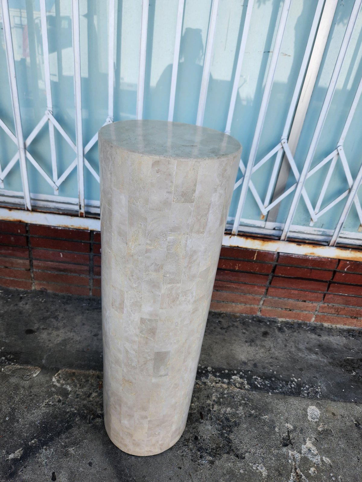Tall cylindrical concrete bollard on a paved surface with blue metal security gate in the background