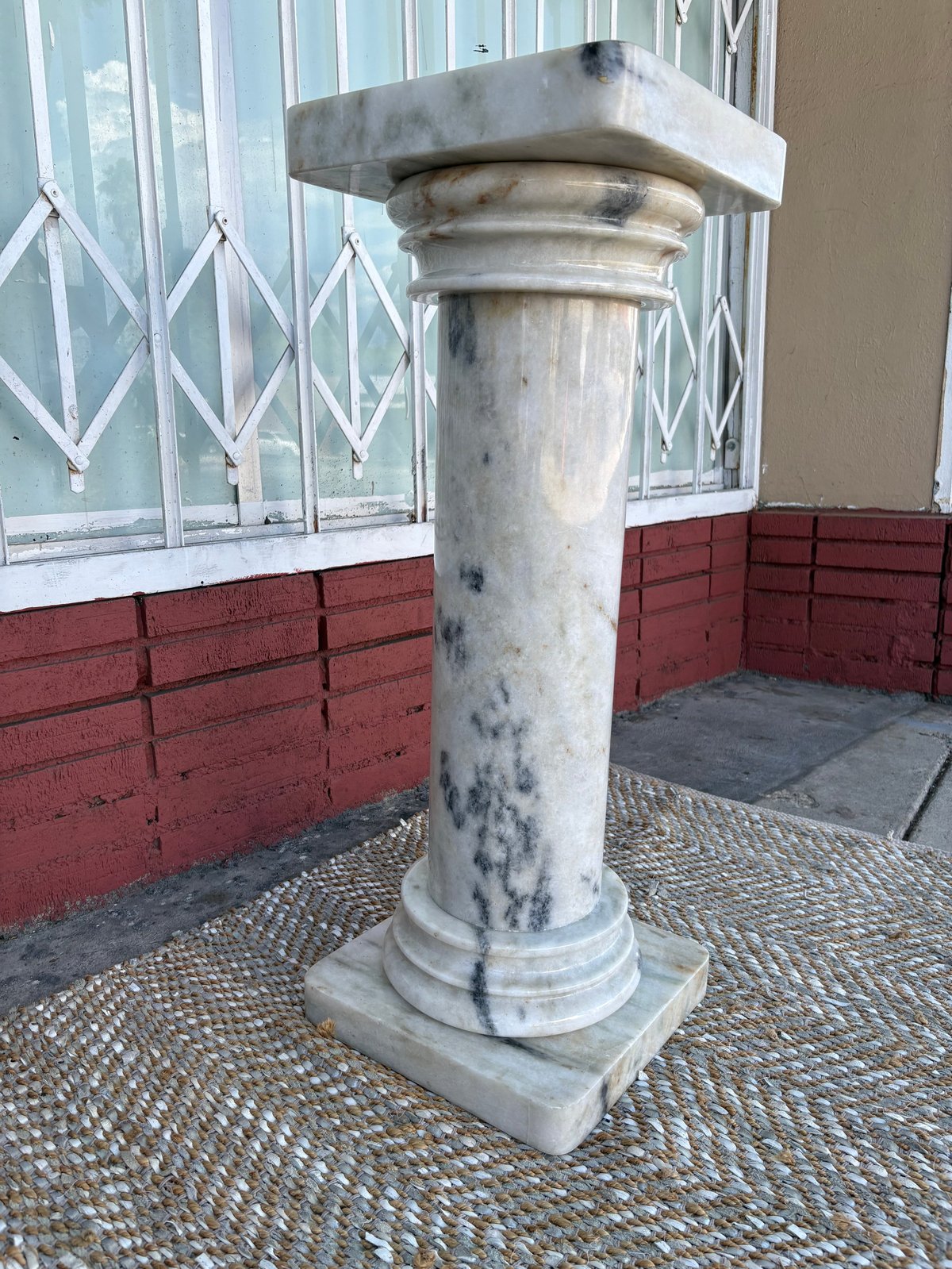 Weathered stone column pedestal with a square capital top on a patterned floor against a red brick wall and white latticed windows