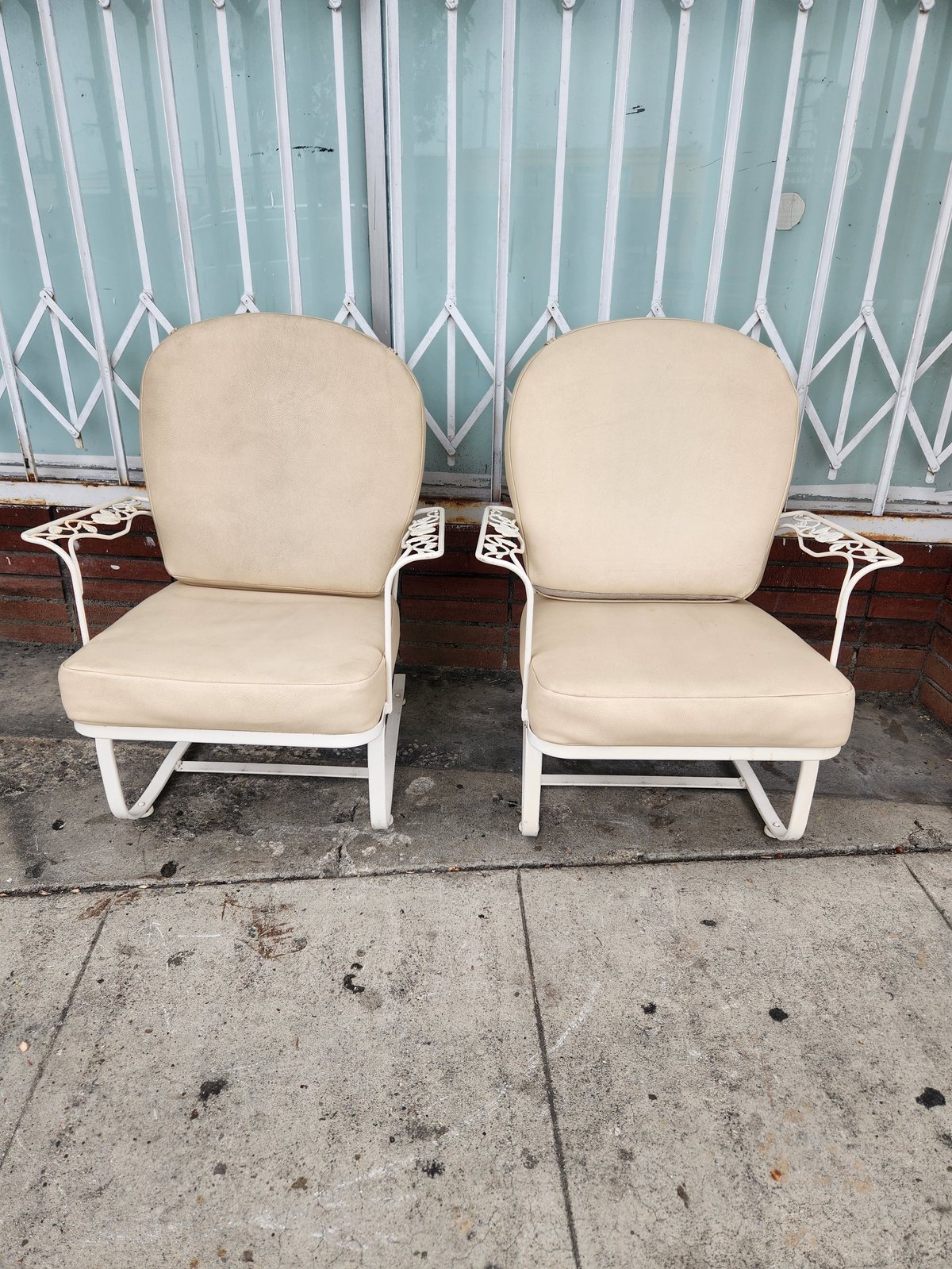 Two cream-colored outdoor patio chairs with white metal frames sitting on concrete ground against a turquoise corrugated metal gate