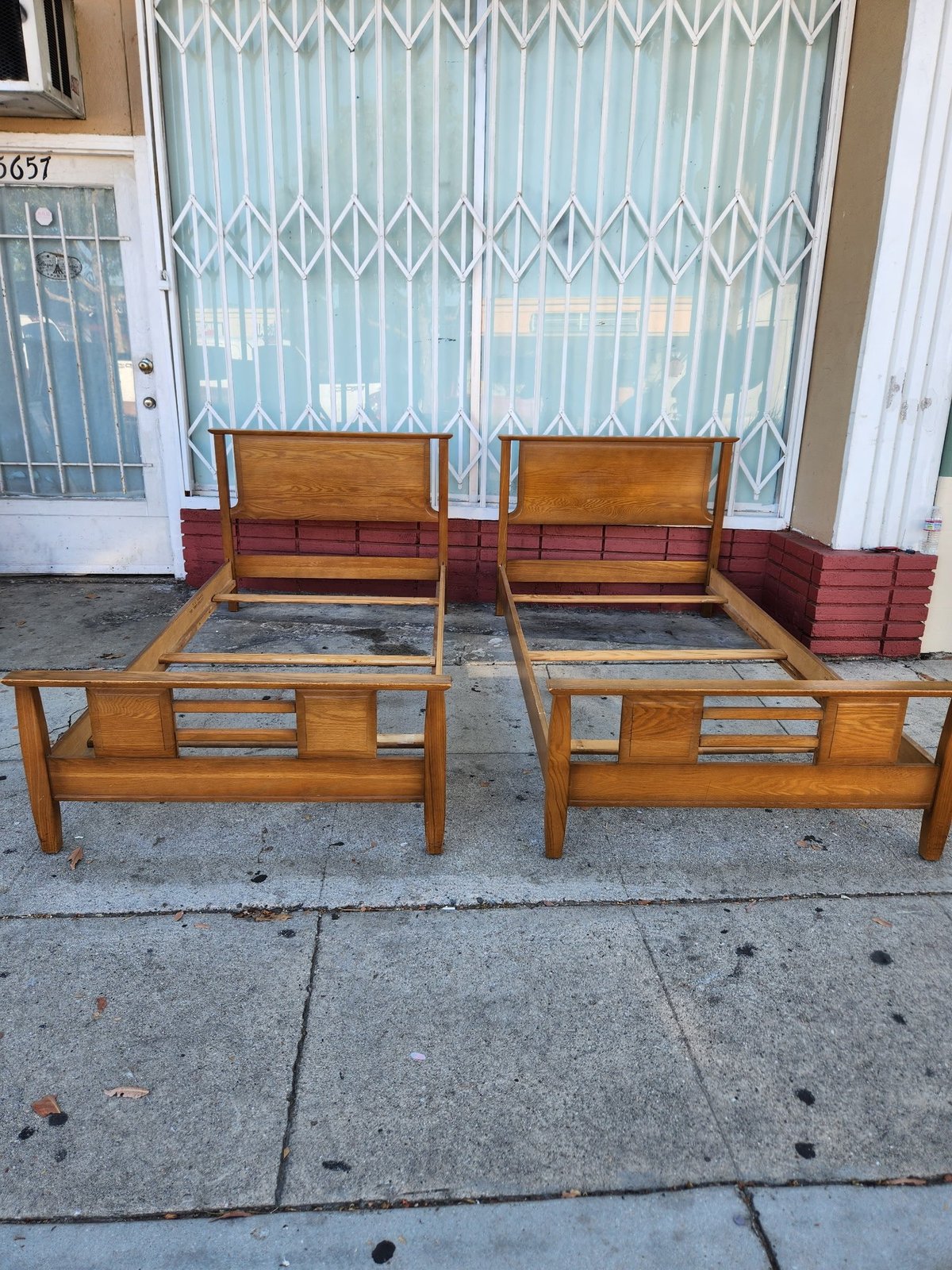 Two wooden twin beds without mattresses positioned on a driveway in front of a white house with metal security gate